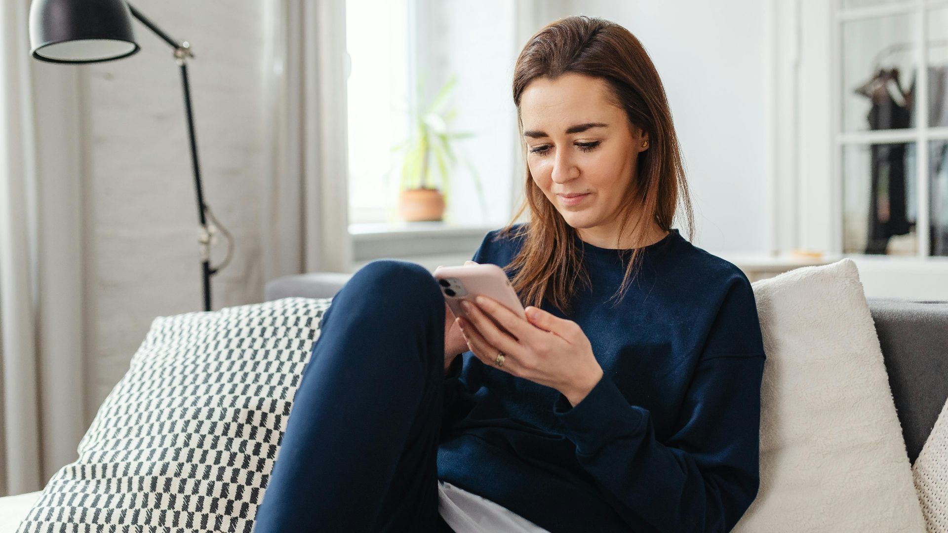 Woman in a blue sweater using a smartphone indoors, relaxing on the couch.