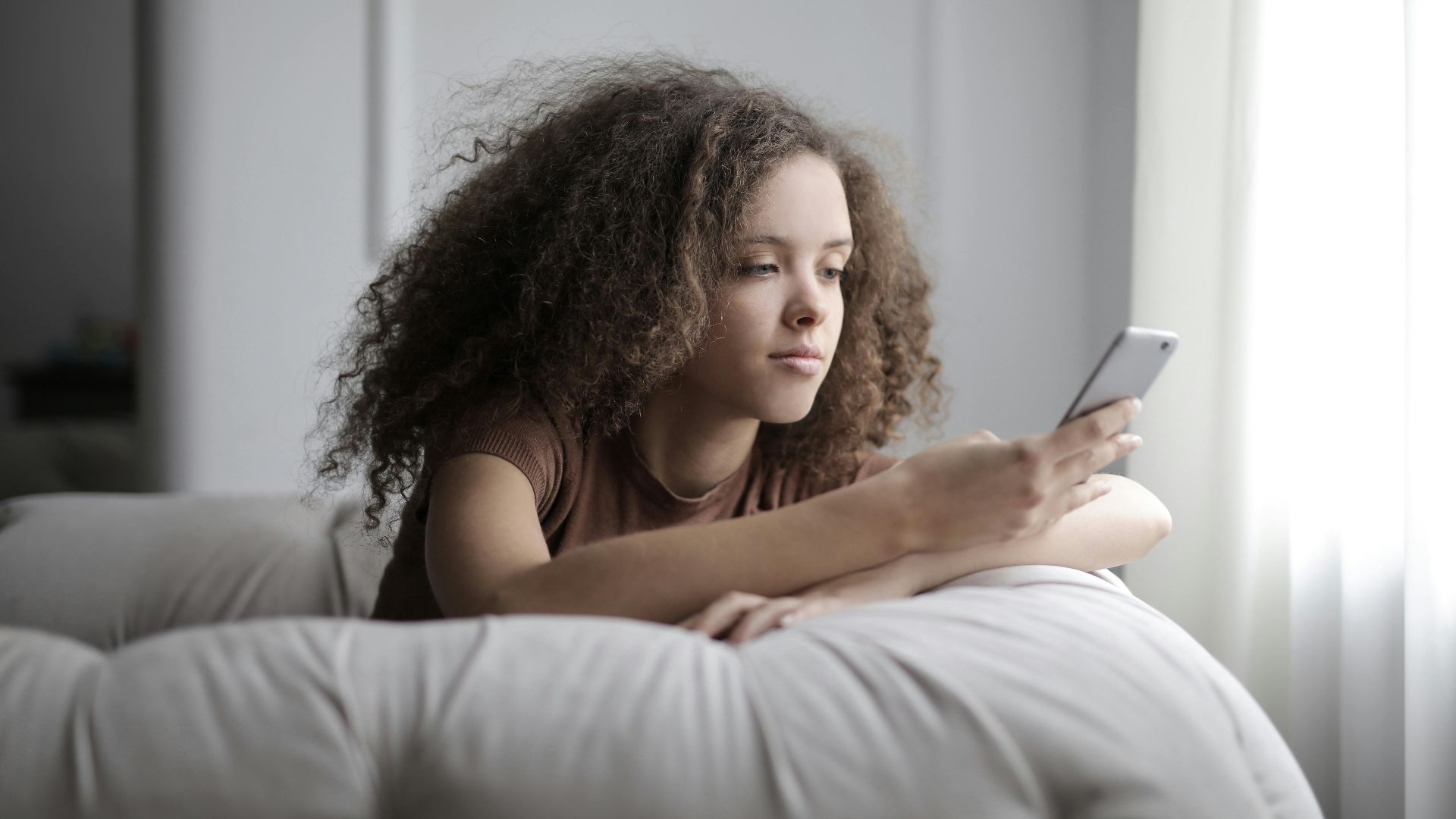 A young woman with curly hair comfortably using her smartphone while sitting indoors.
