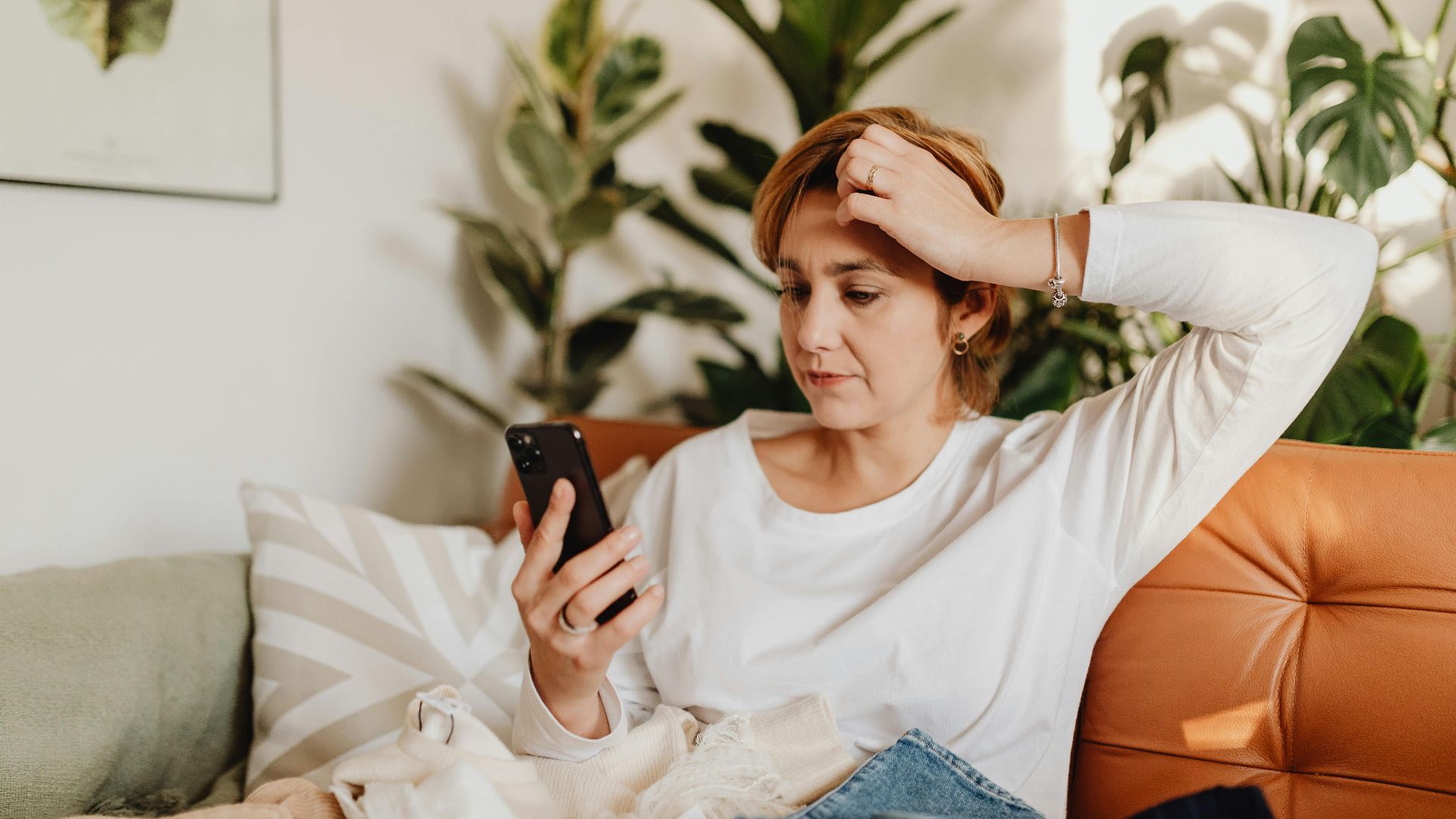 Young woman in white blouse using smartphone while sitting on an orange sofa surrounded by plants.