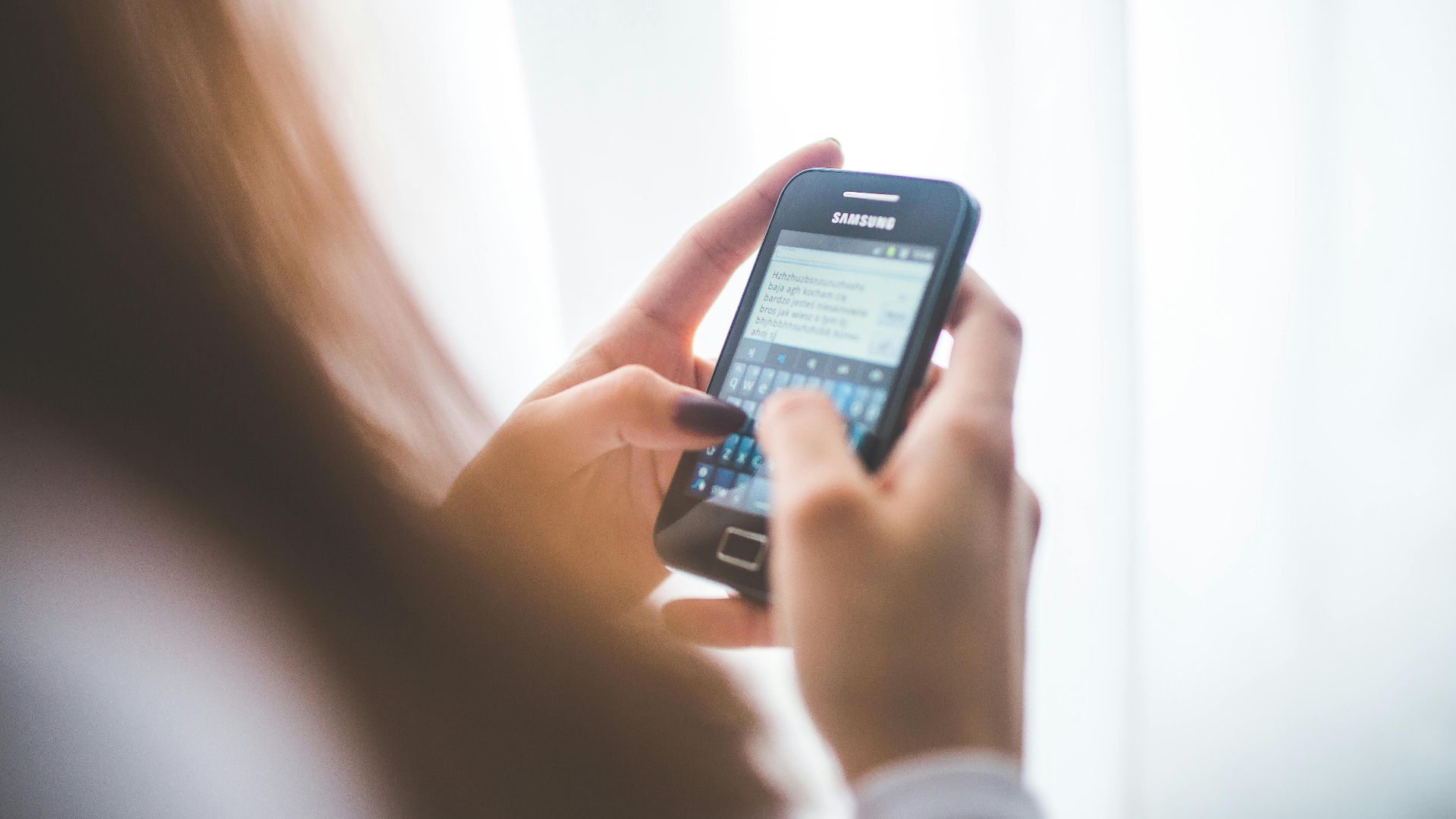 Close-up of a young woman typing on a Samsung smartphone at home. Bright indoor lighting.