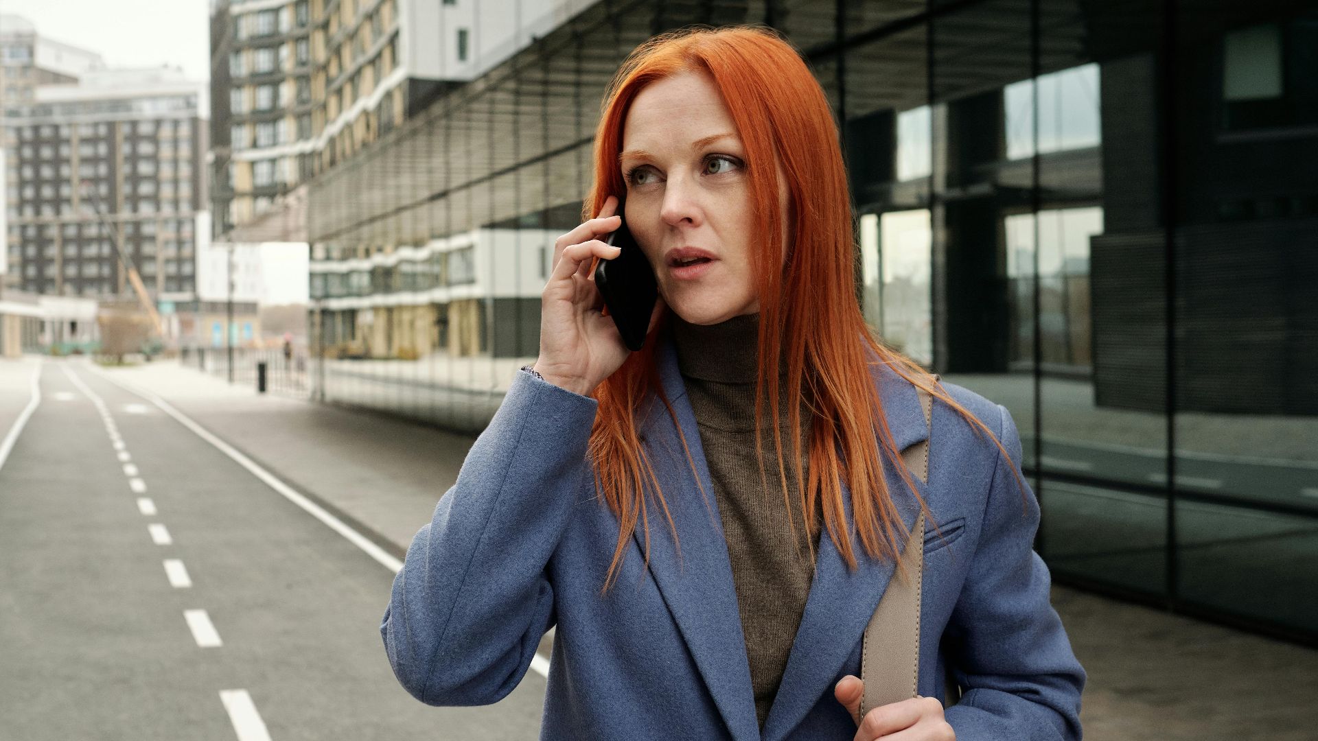 Red-haired woman in blue coat making a phone call on an empty city street.