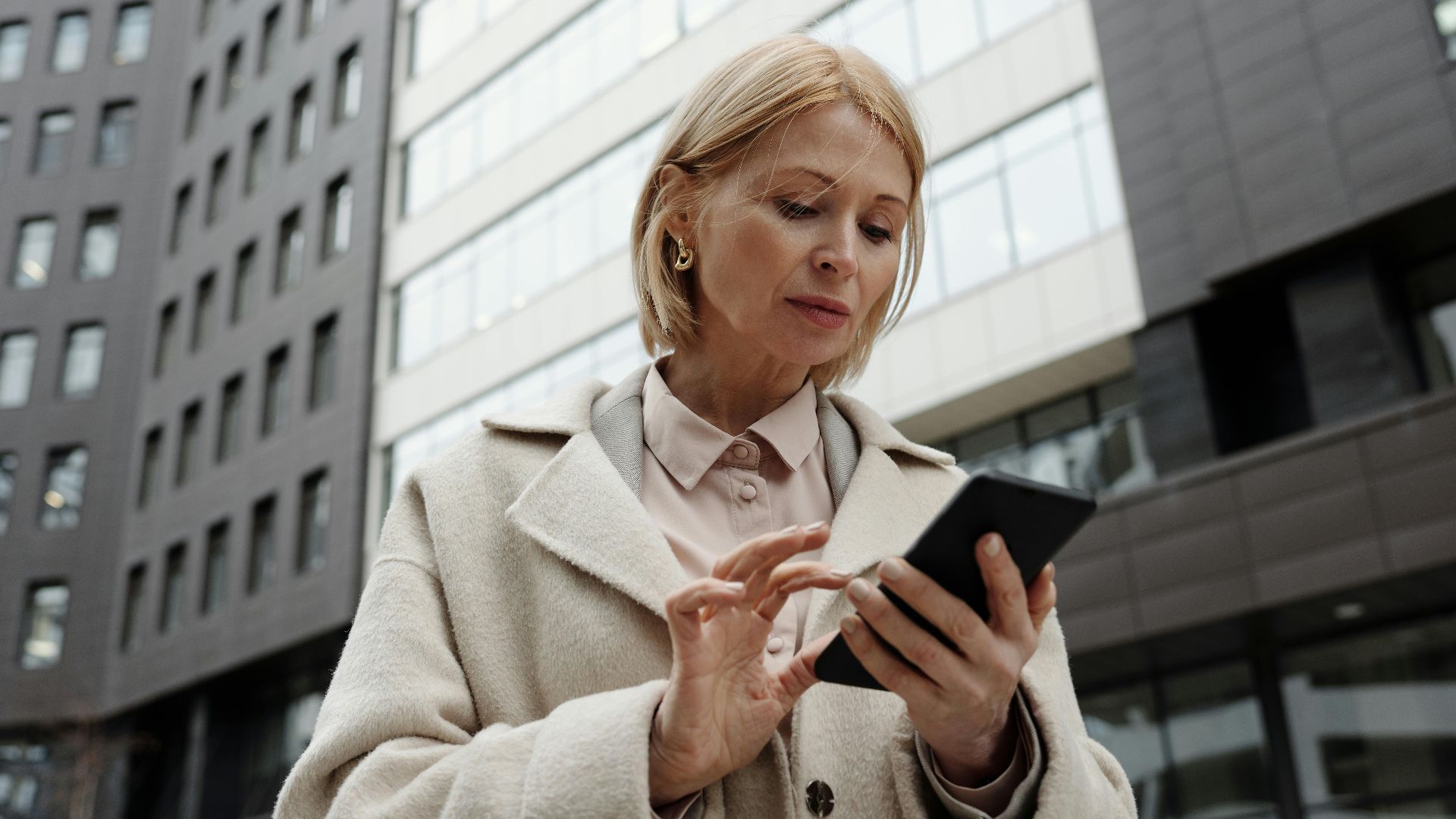 Woman in coat using smartphone in front of modern building. Professional and focused expression.