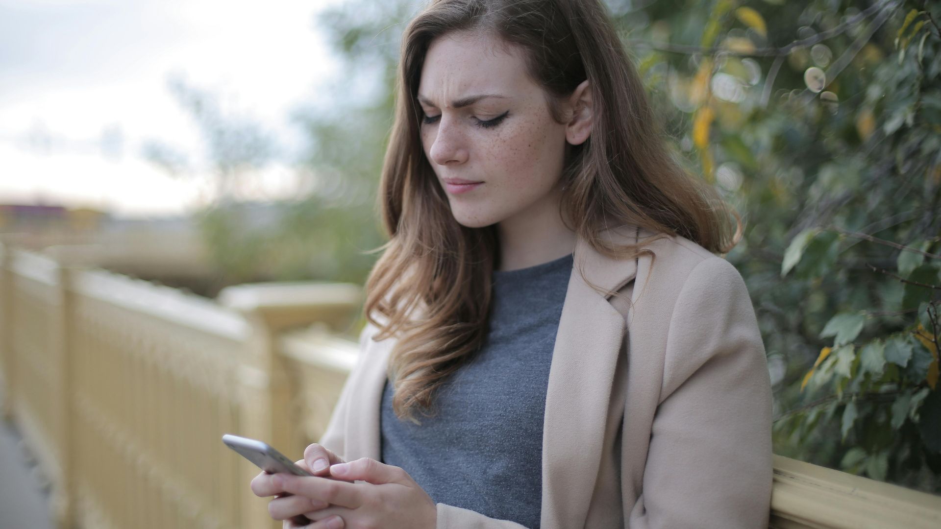 A woman stands outdoors in a casual outfit, using her smartphone with a pensive look.