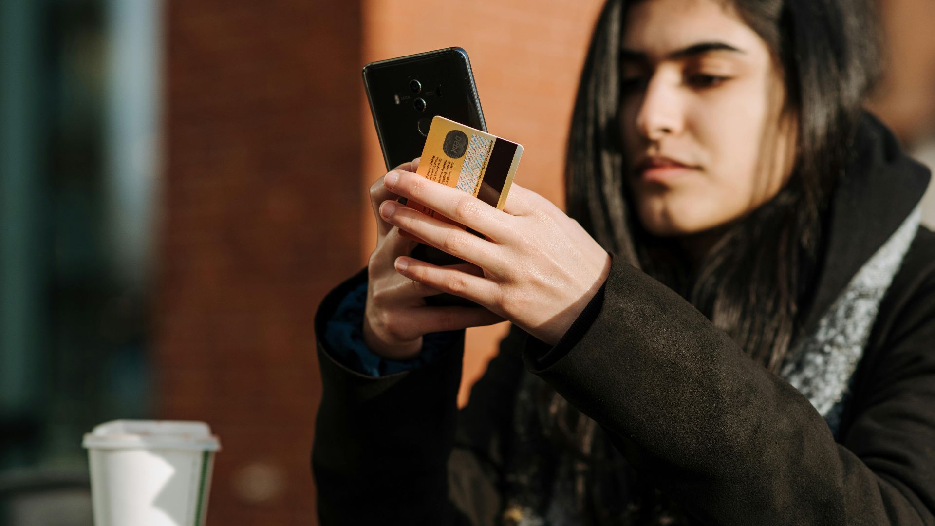 Crop young Hispanic female shopper with debit card making purchase on cellphone near takeaway beverage in cafeteria