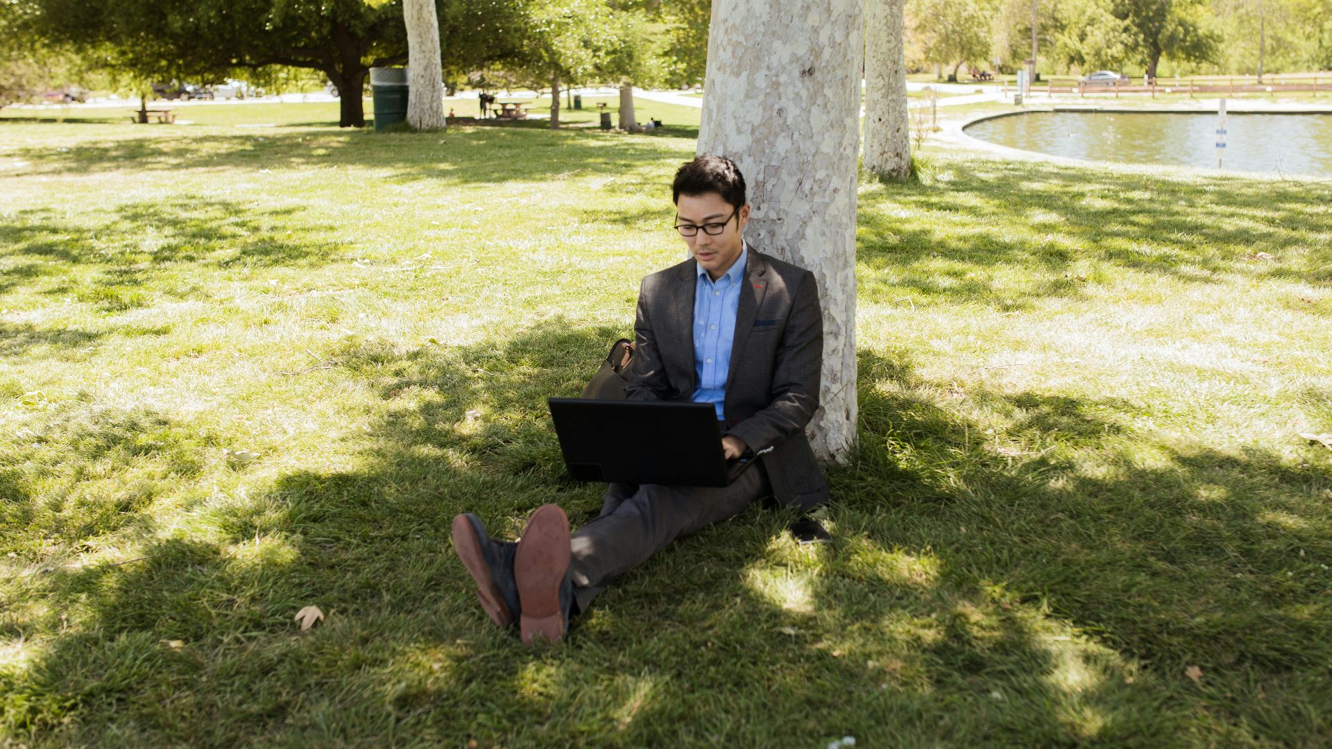 Businessman in a suit using a laptop under a tree in a sunny park, engaging in remote work.