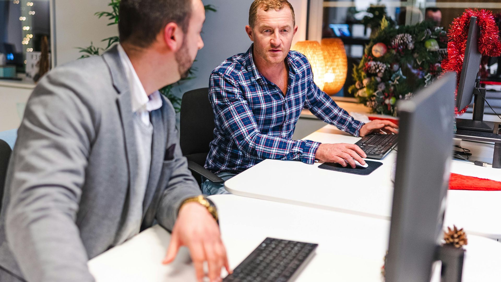 Two men collaborate in a modern office with computers and holiday decor.