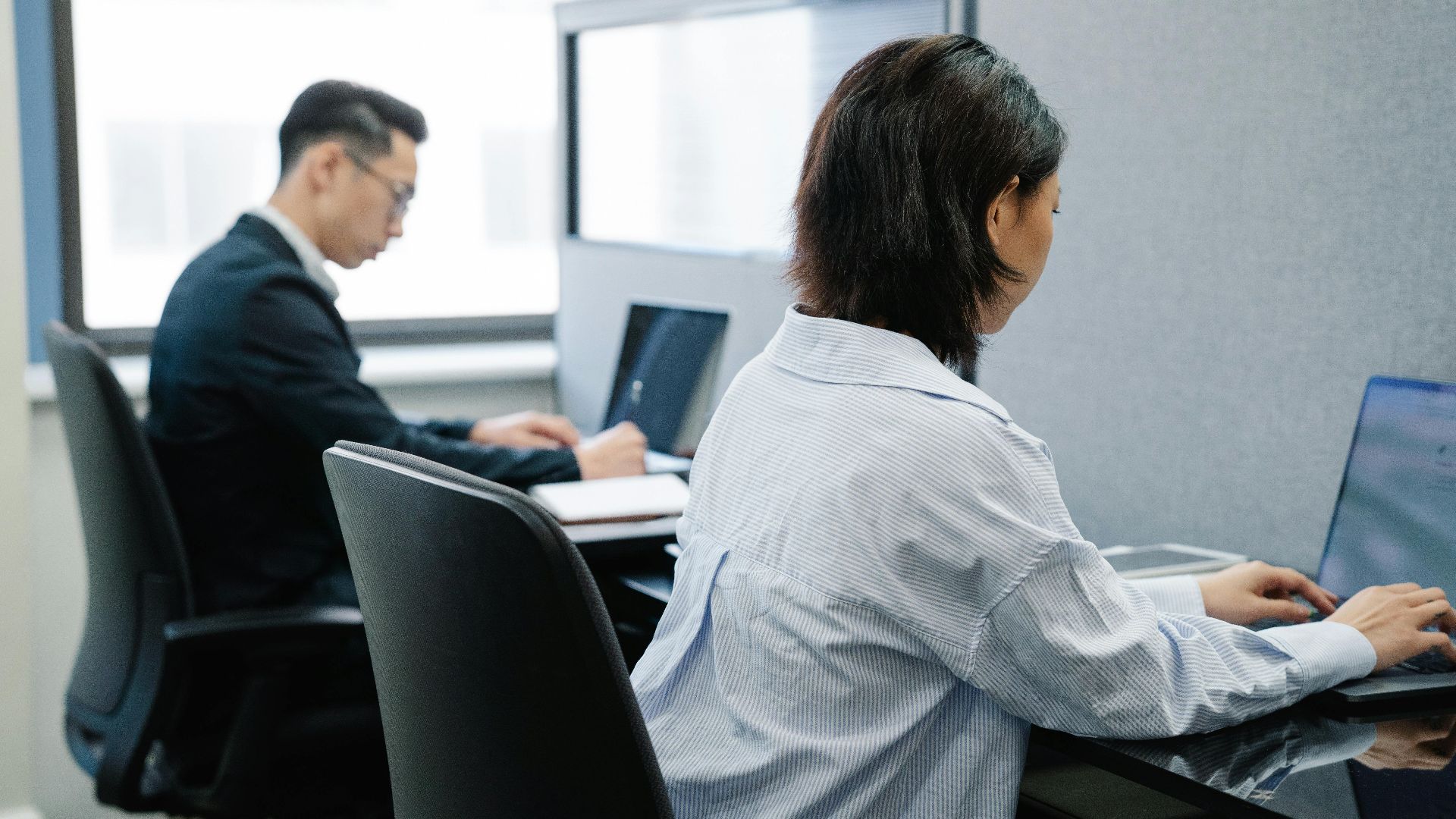 Two professionals working on laptops in a modern office cubicle setup.