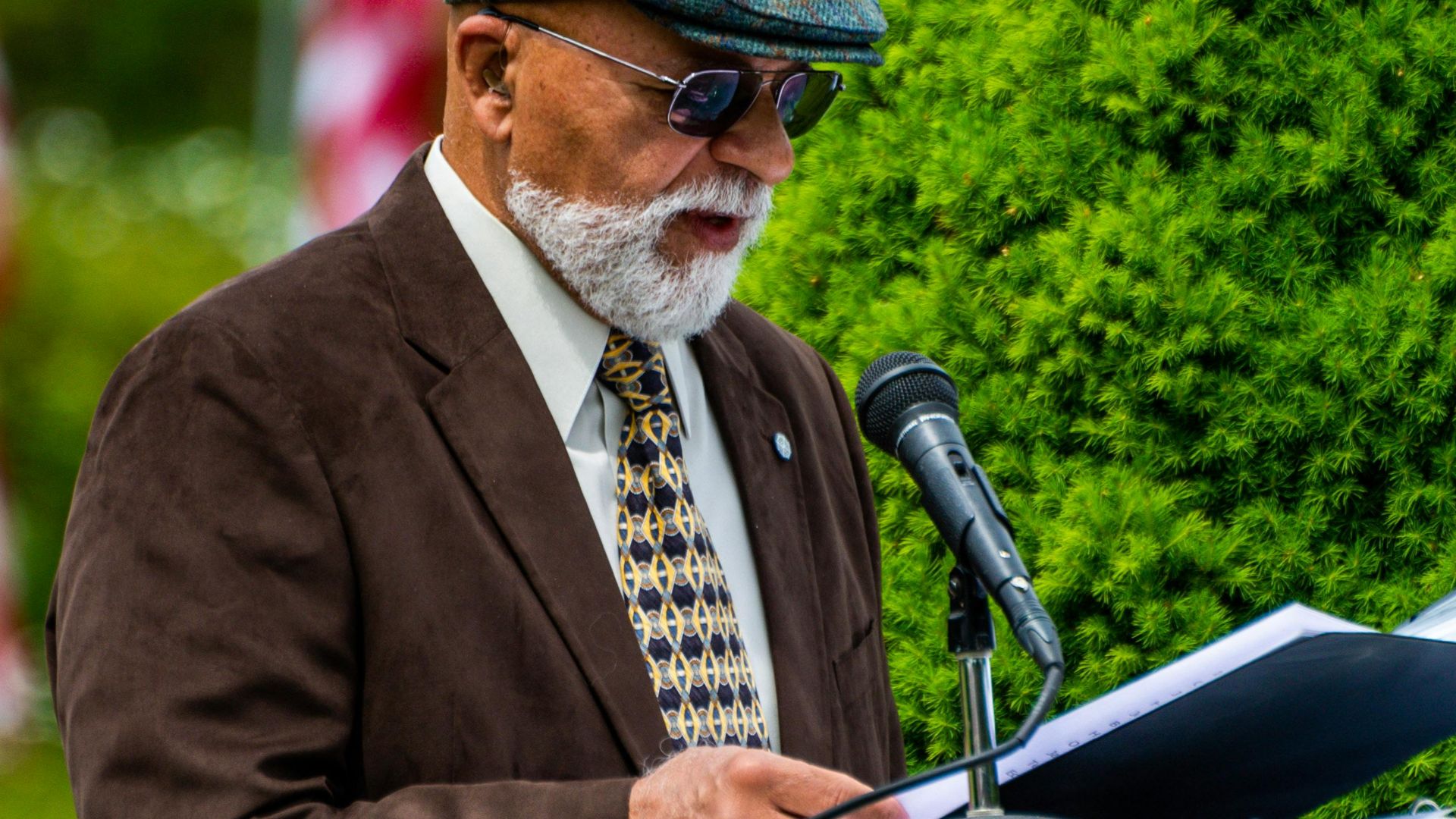 Elderly man in brown suit speaking outdoors on a sunny day.