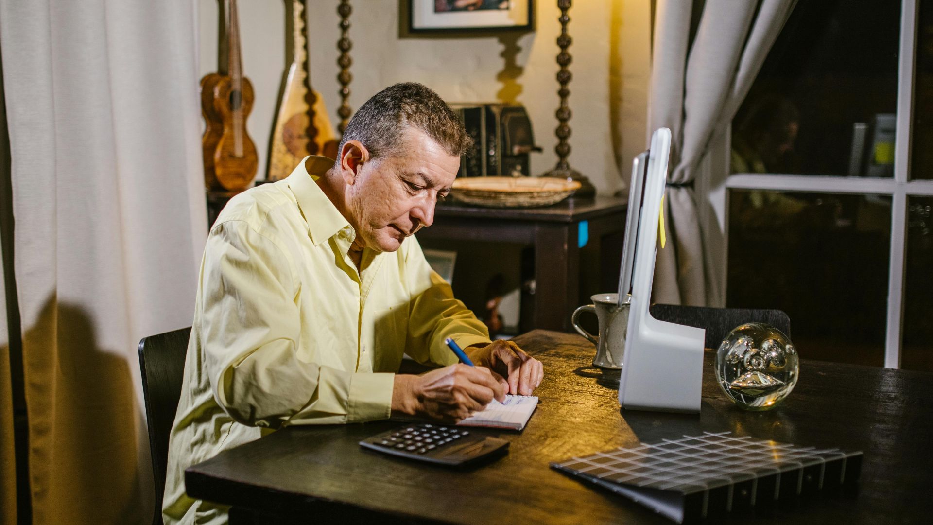 Senior man wearing yellow shirt writing notes at desk with lamp, calculator, and glass sphere nearby.