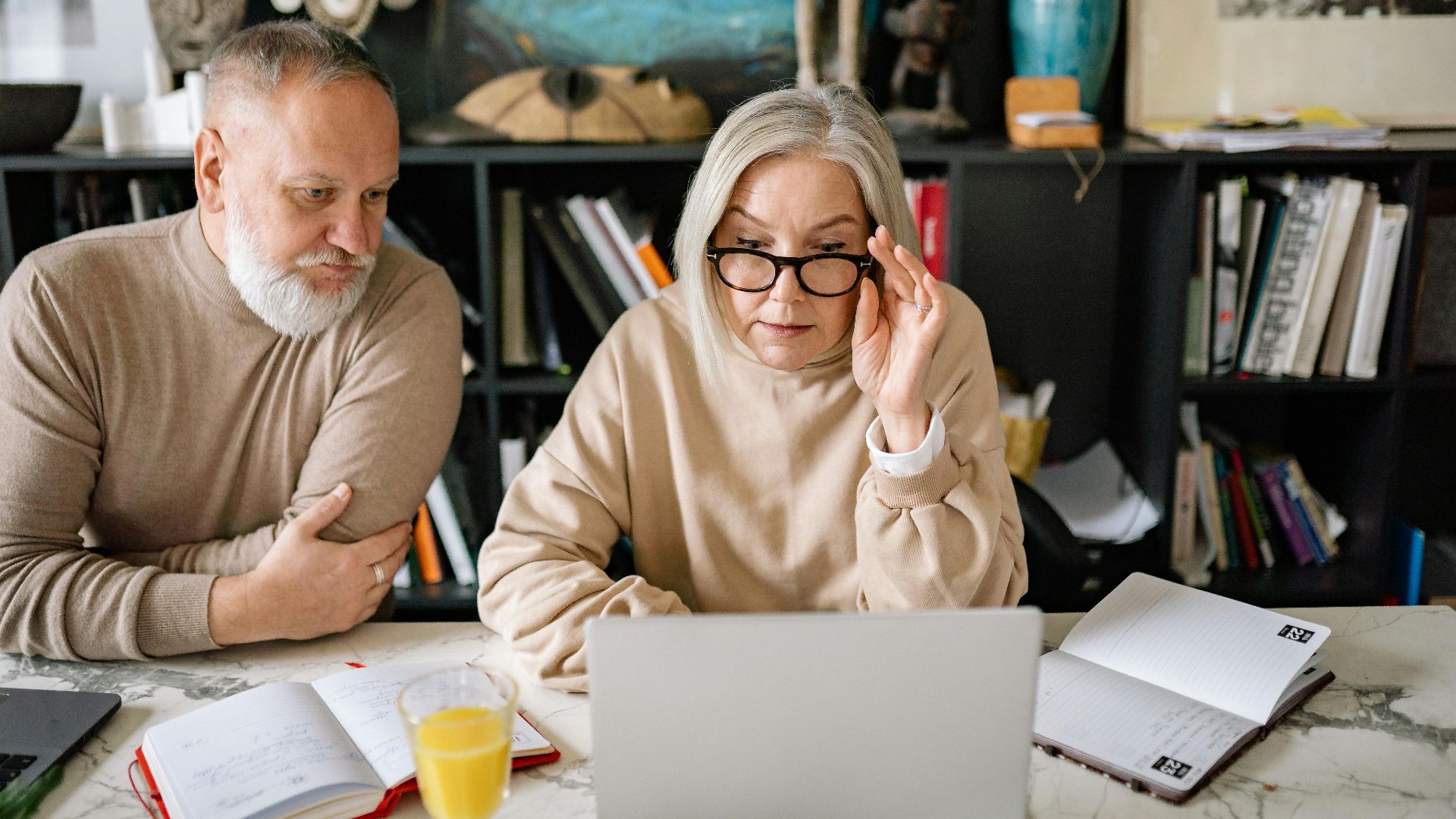 An elderly couple in a cozy home setting engaged with a laptop, planners, and a glass of juice on the table.