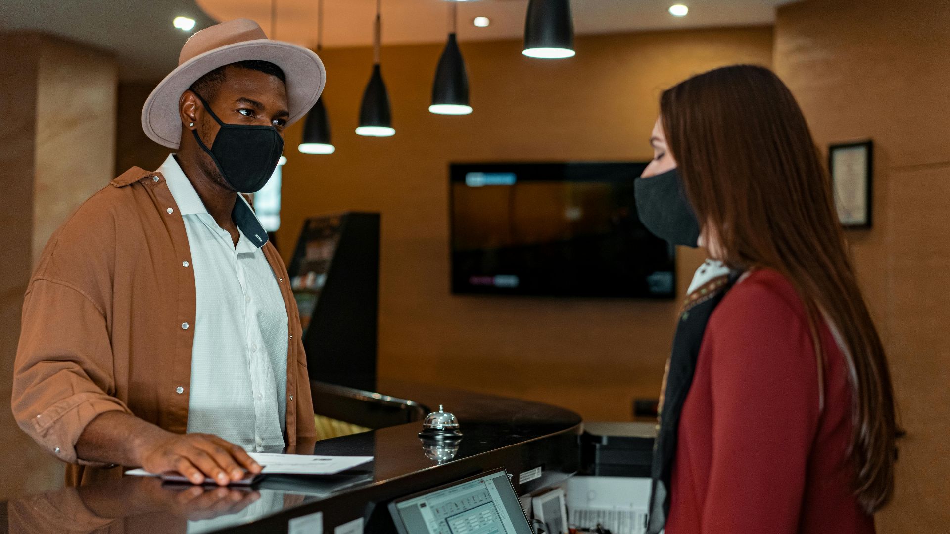A man and woman in face masks checking in at a hotel reception, emphasizing safety protocols.