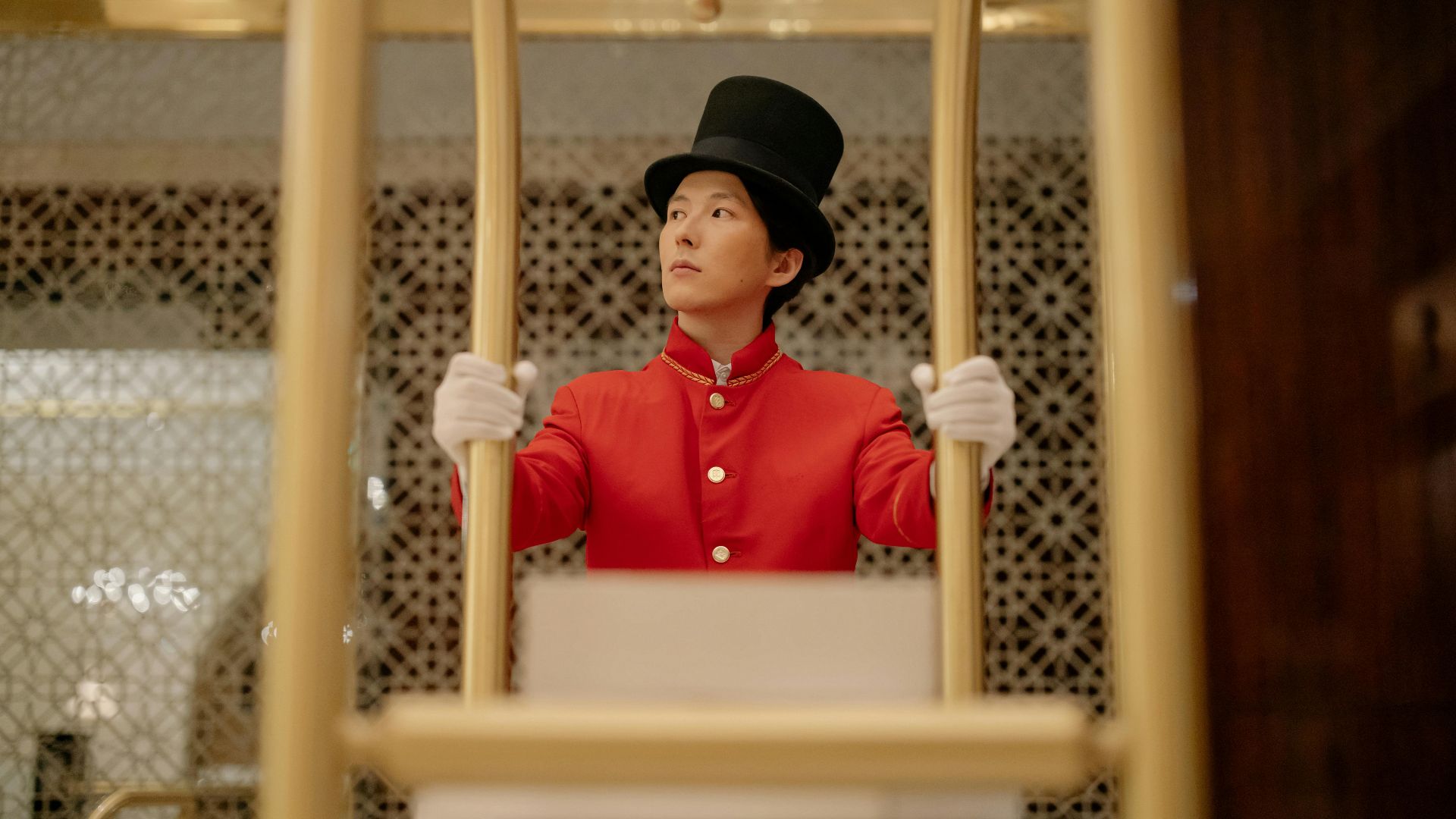 A bellboy in a red uniform stands with a luggage cart inside a luxurious hotel.
