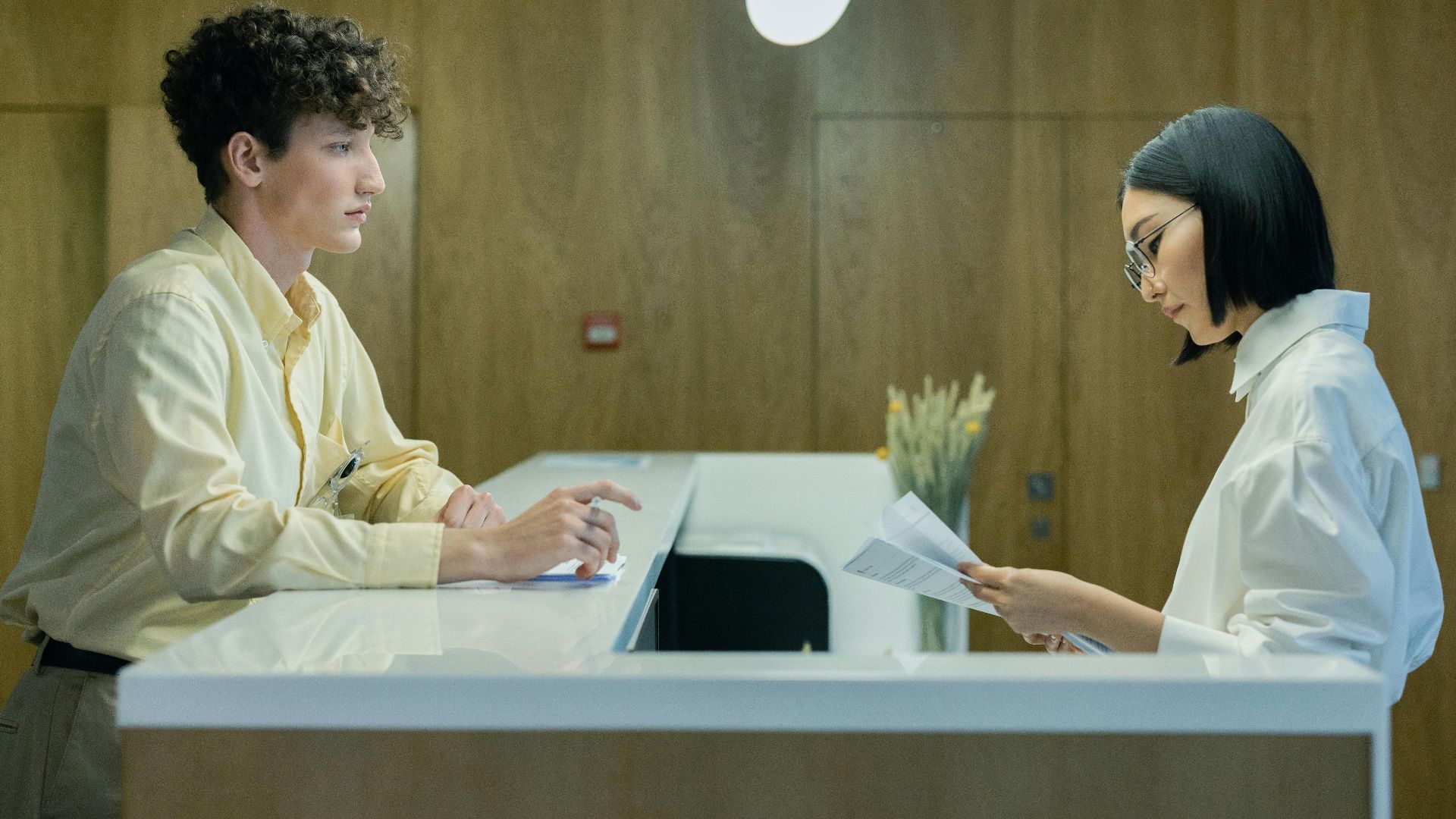 A young man and woman interacting at a modern office reception desk indoors.