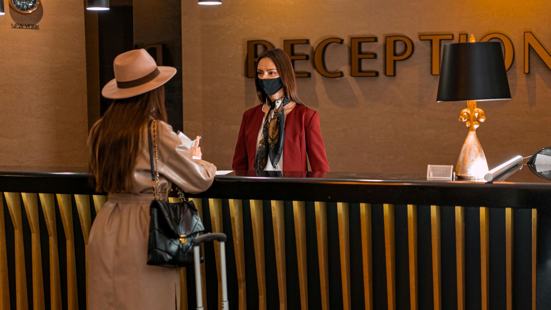 A woman speaks to a receptionist wearing a mask at a hotel reception.