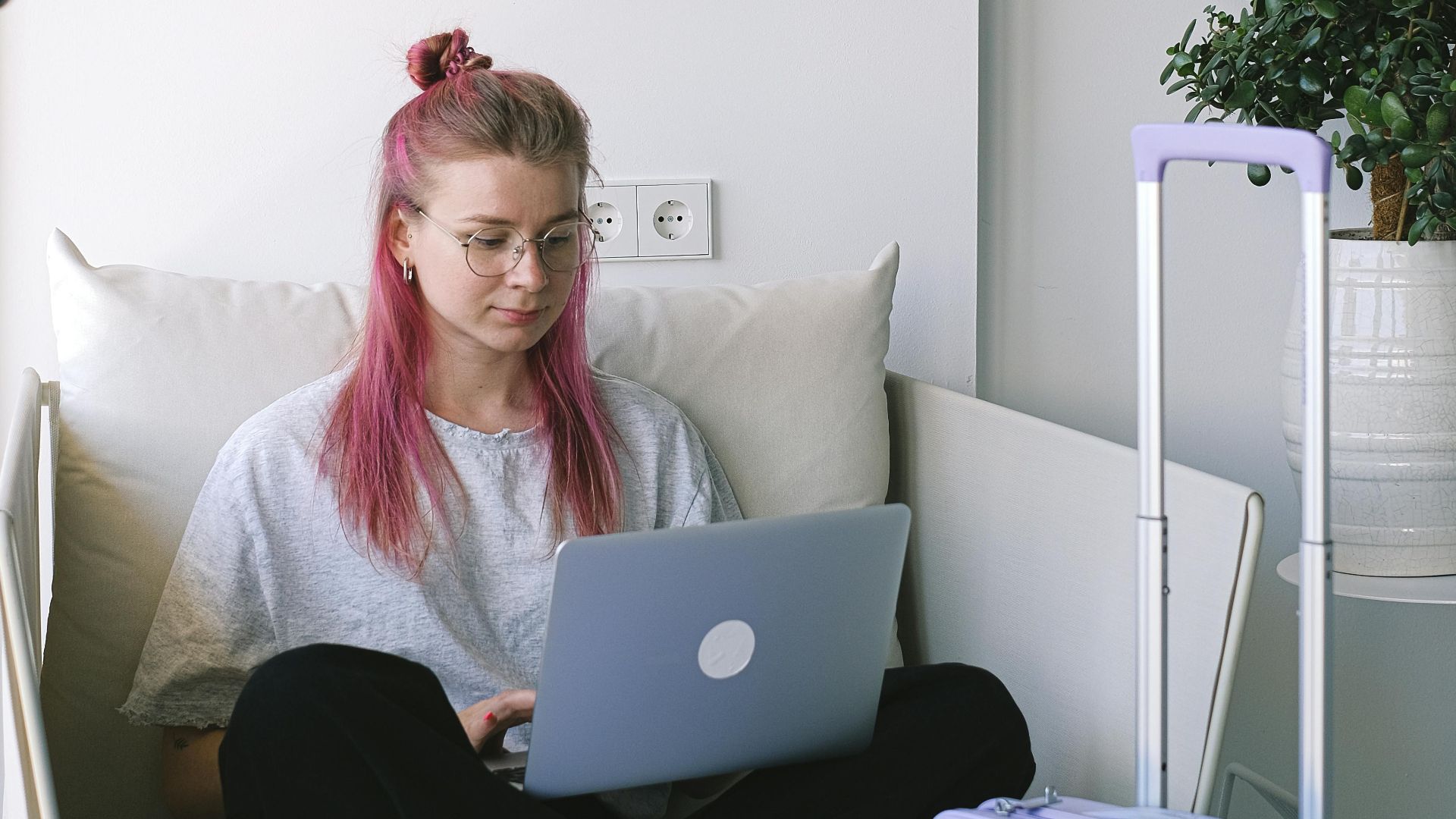Woman with pink hair working on a laptop while sitting on a sofa in a hotel room with luggage.
