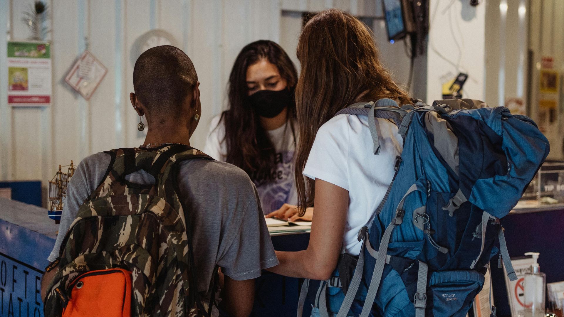 Two backpackers checking in at a hostel reception, wearing face masks.