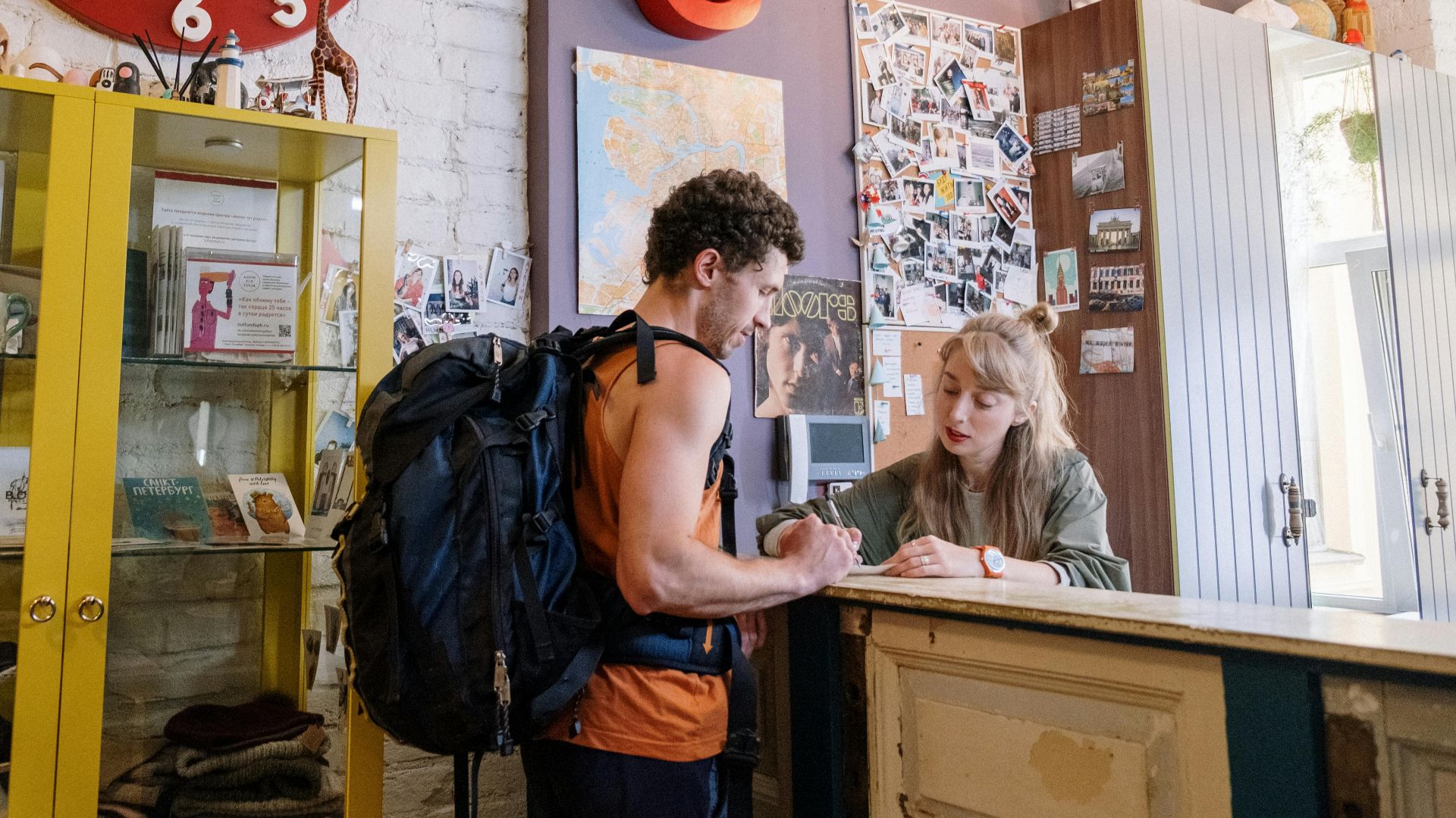 A backpacker at a hostel front desk getting assistance from a receptionist.