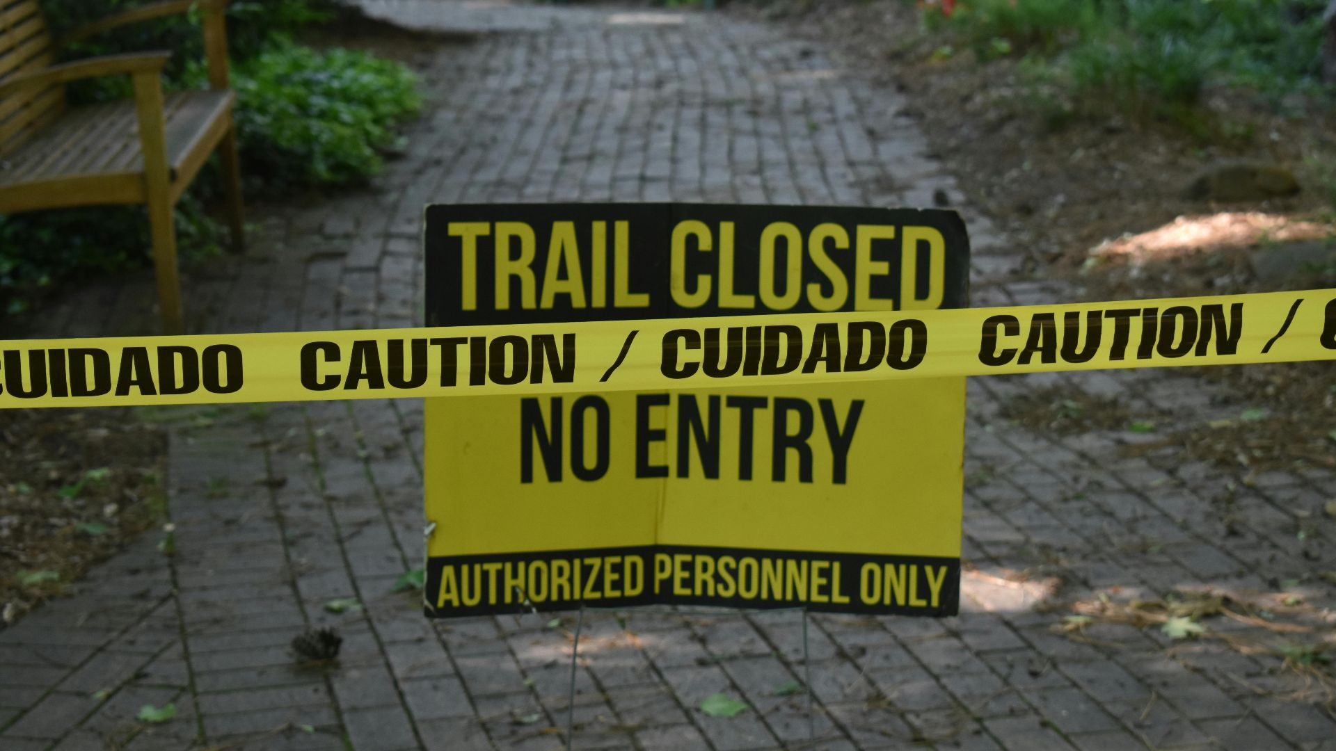Trail closed sign with caution tape blocking pathway in a park setting.