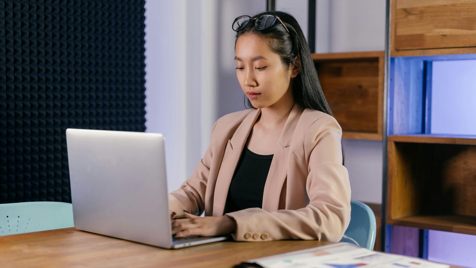 Asian businesswoman in a blazer typing on a laptop in a modern office setting.