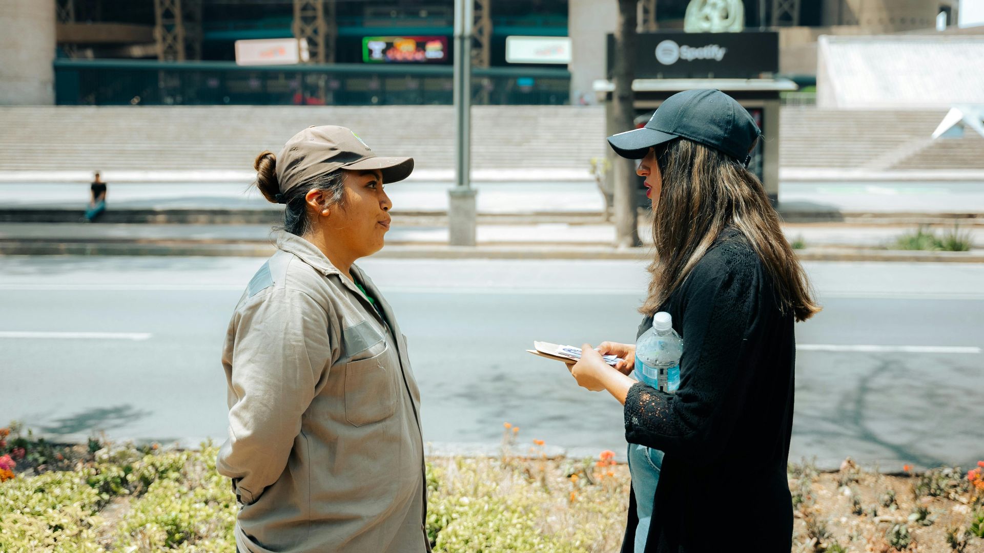 Two women in conversation on an urban street with a modern building backdrop.