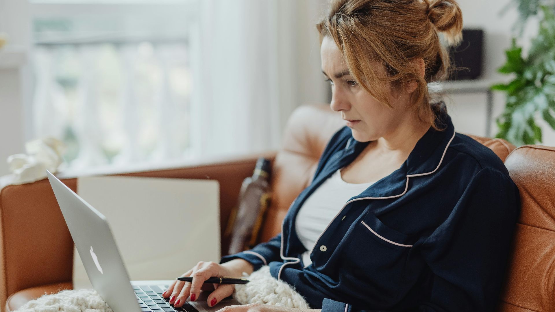 Woman in pajamas working on a laptop from a cozy home environment, focused on her task.