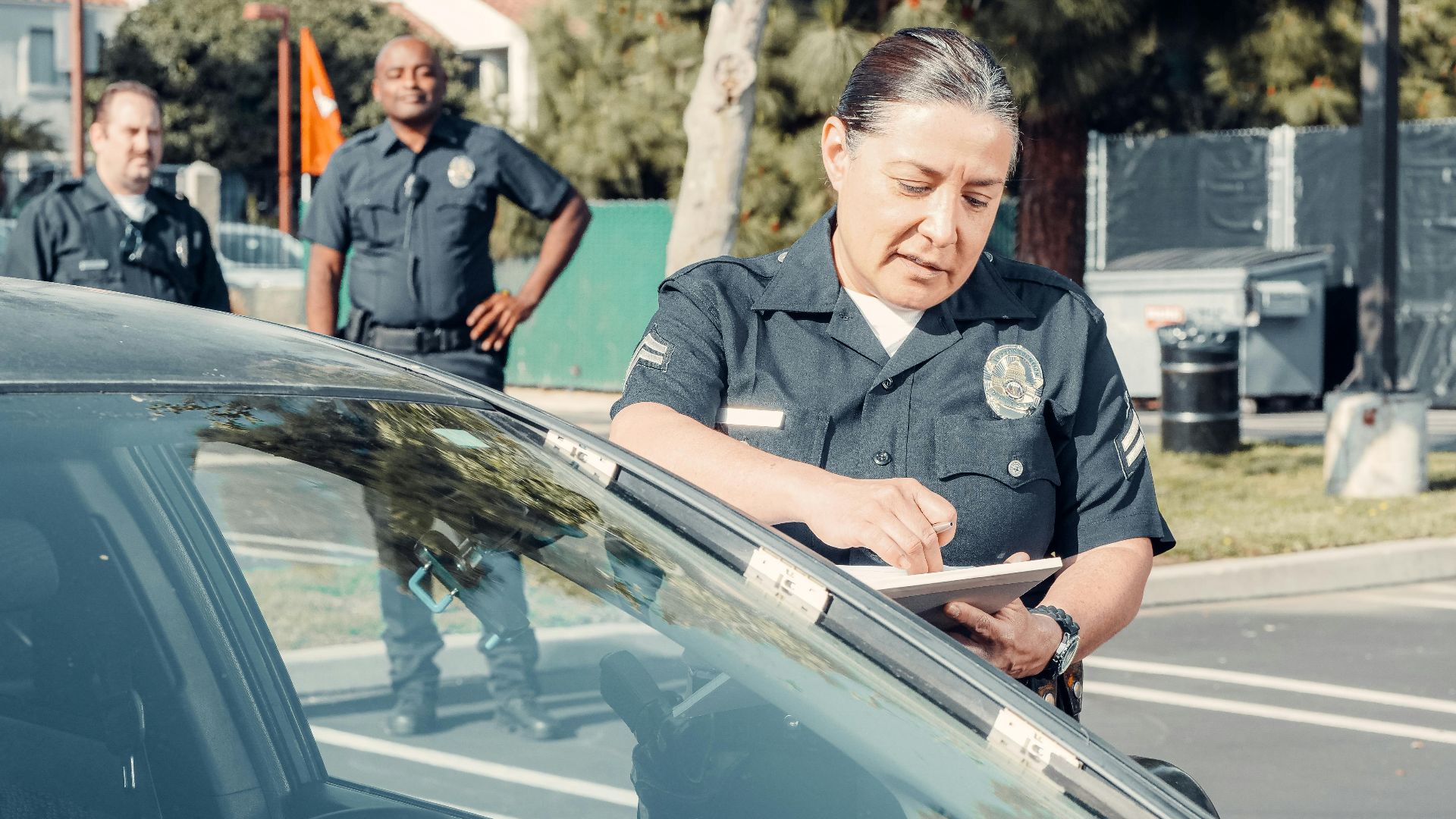 Police officers handling a traffic violation in a parking lot, focus on policewoman writing a ticket.