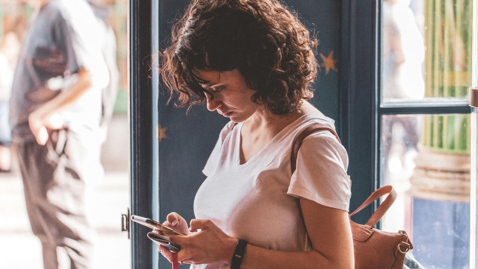 Woman with curly hair using smartphone by a sunlit window, wearing a white t-shirt.