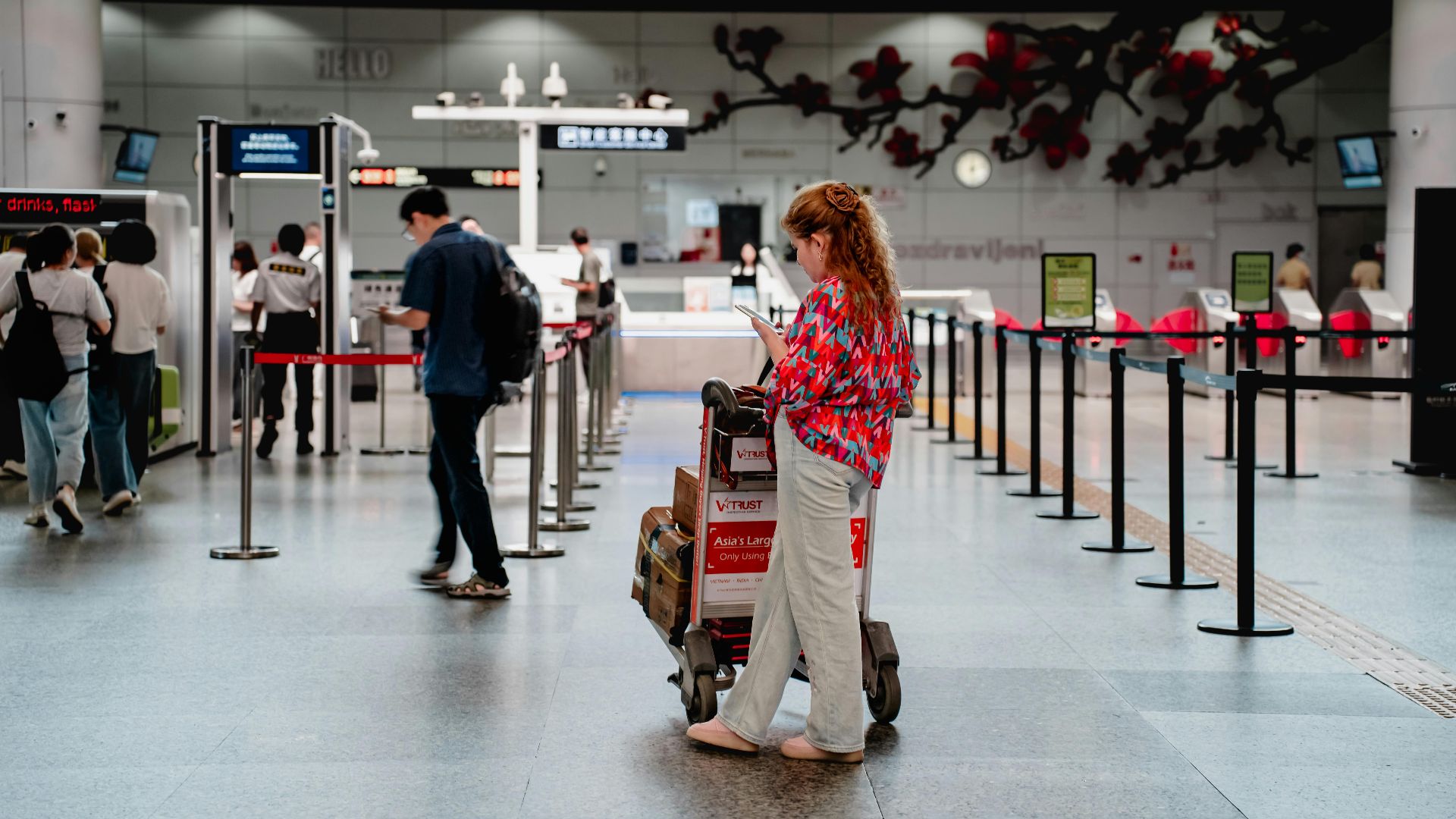 A traveler checks her phone while waiting with a luggage cart at a busy airport terminal in Guangzhou.