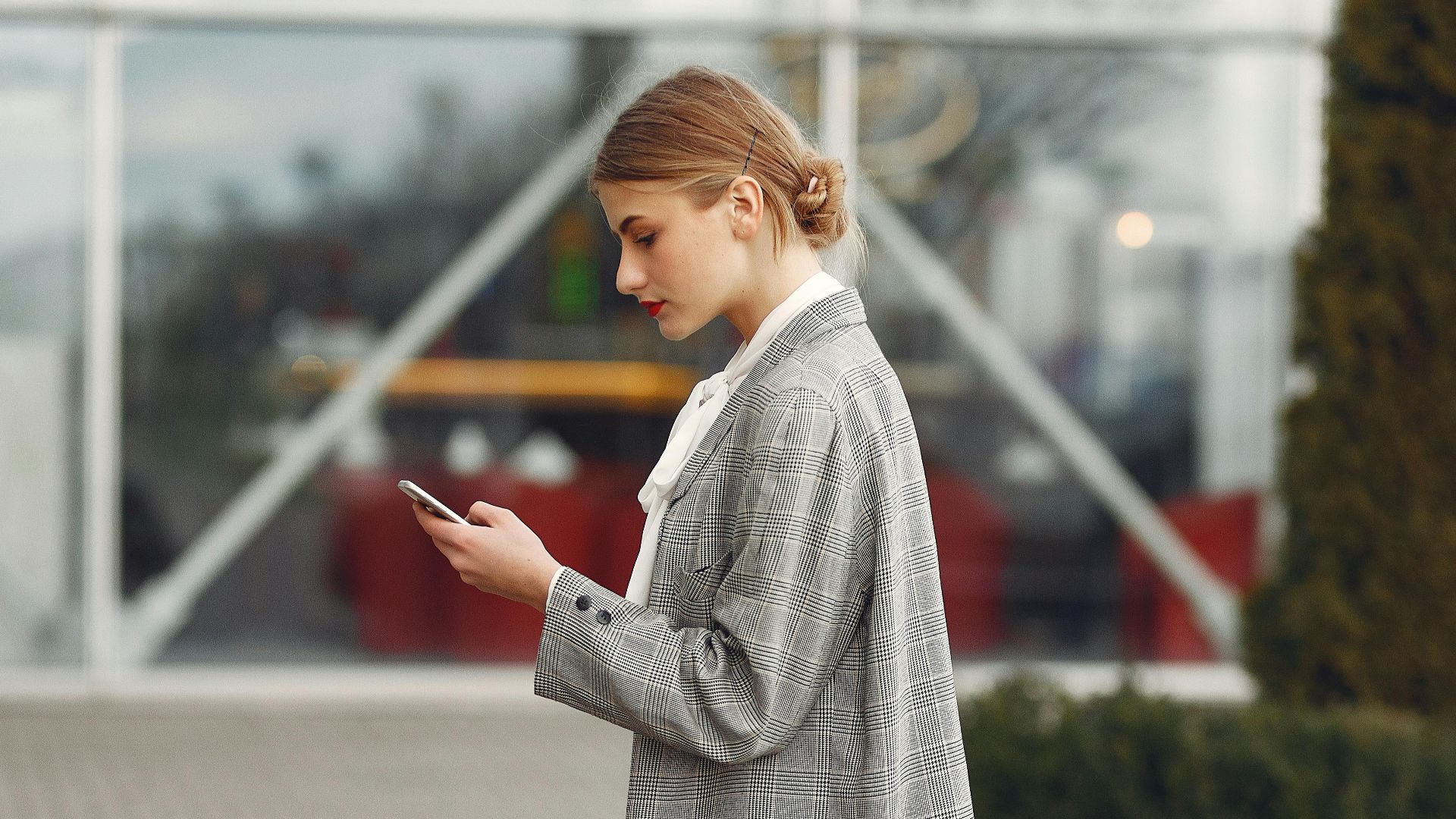 Stylish woman walking with suitcase while using smartphone outside a modern building.