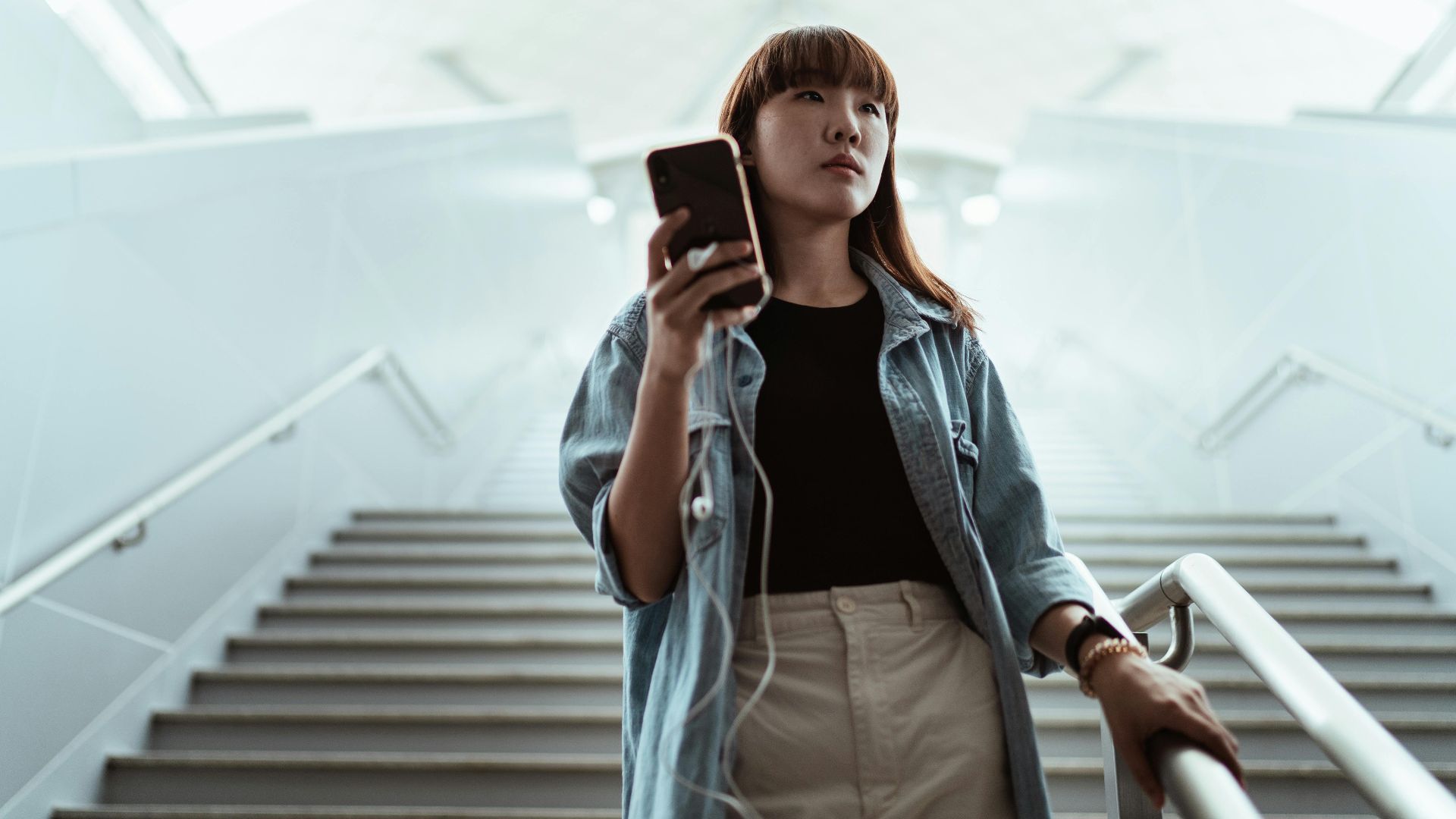 From below of focused young Asian female in stylish wear browsing mobile phone while standing on staircase near railing in spacious light contemporary building