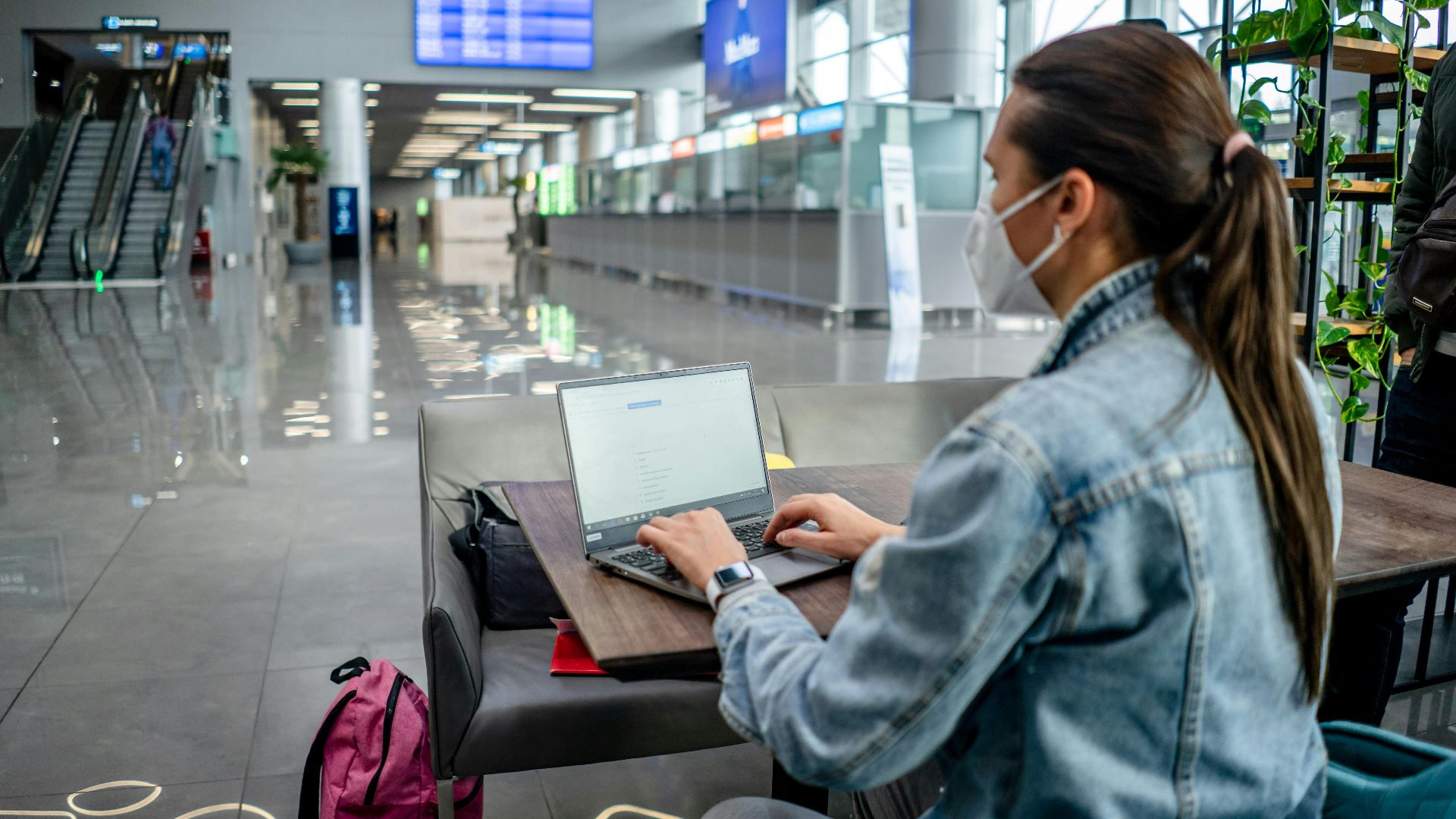 Side view of concentrated female freelancer in protective mask typing on netbook while sitting at table in modern airport terminal