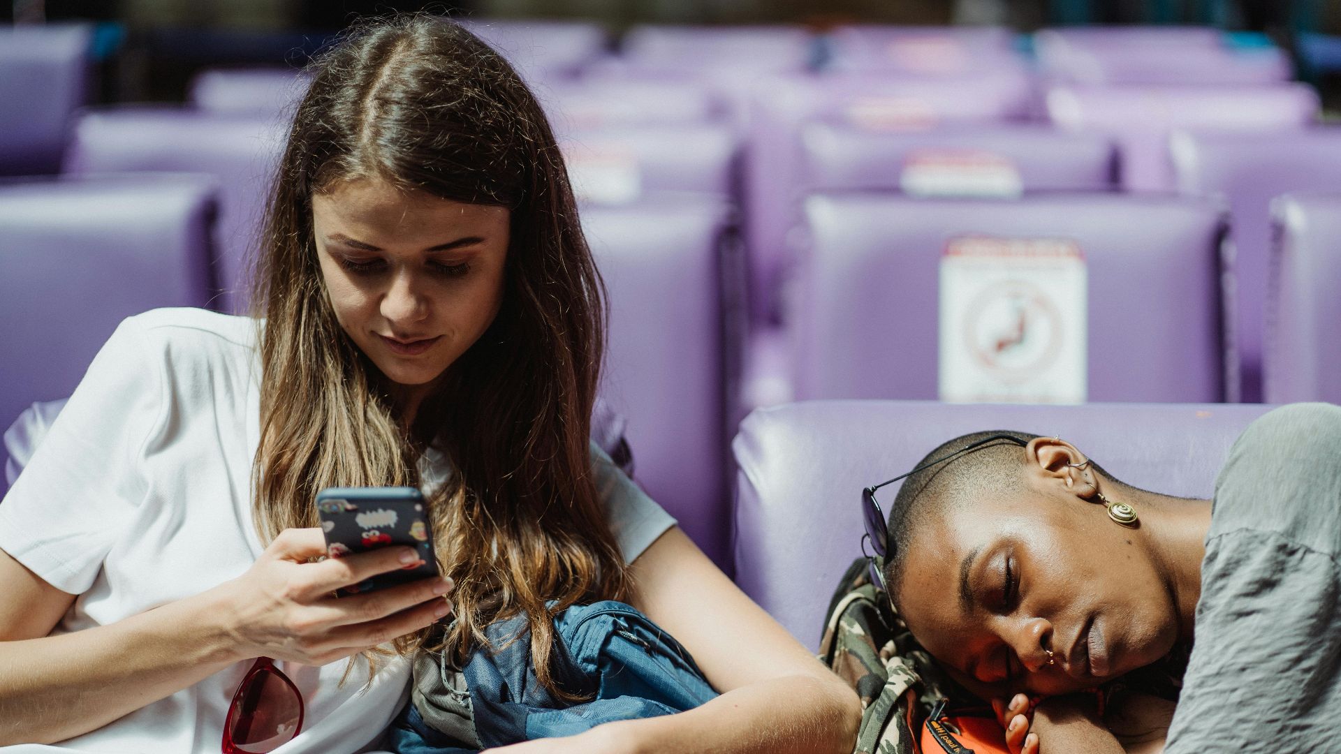Two young women relaxing in a station. One is texting while the other is resting.
