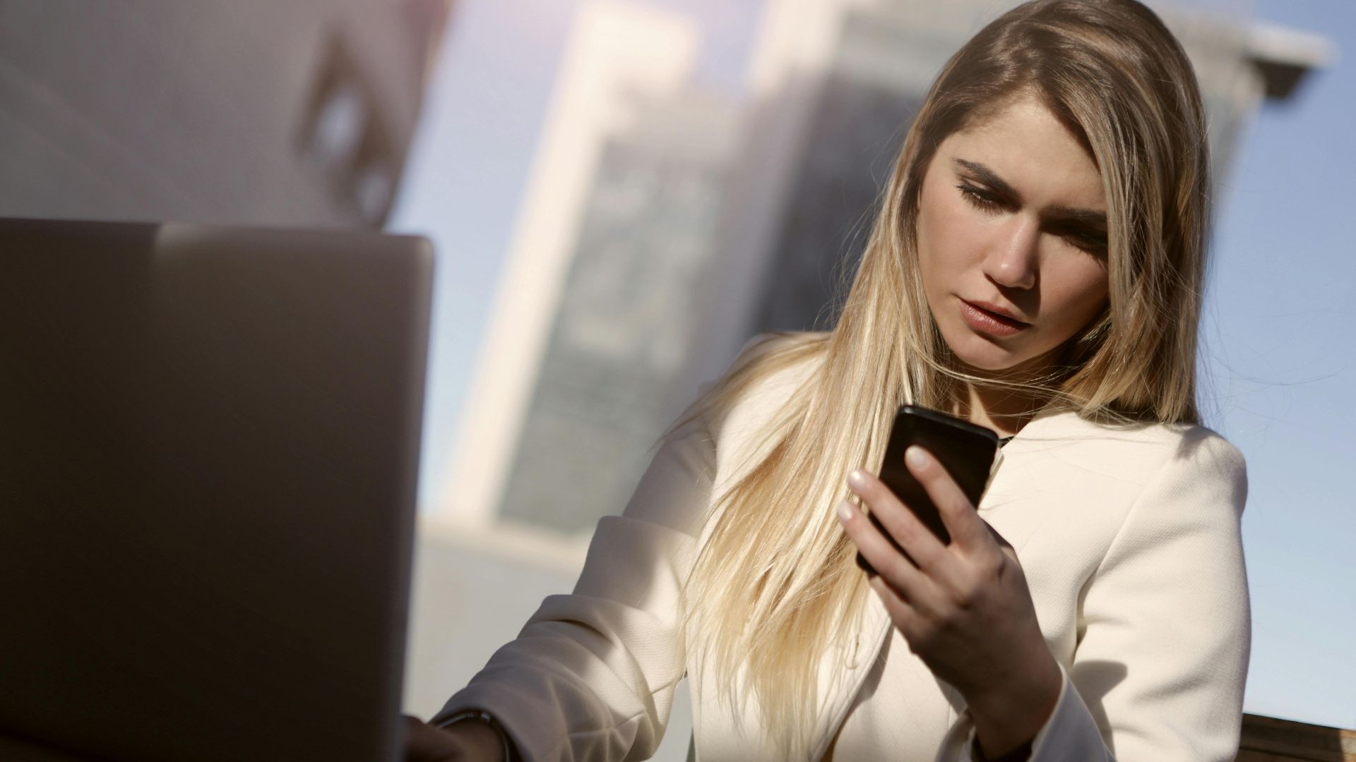 Focused businesswoman using a smartphone outdoors with laptop, modern lifestyle.