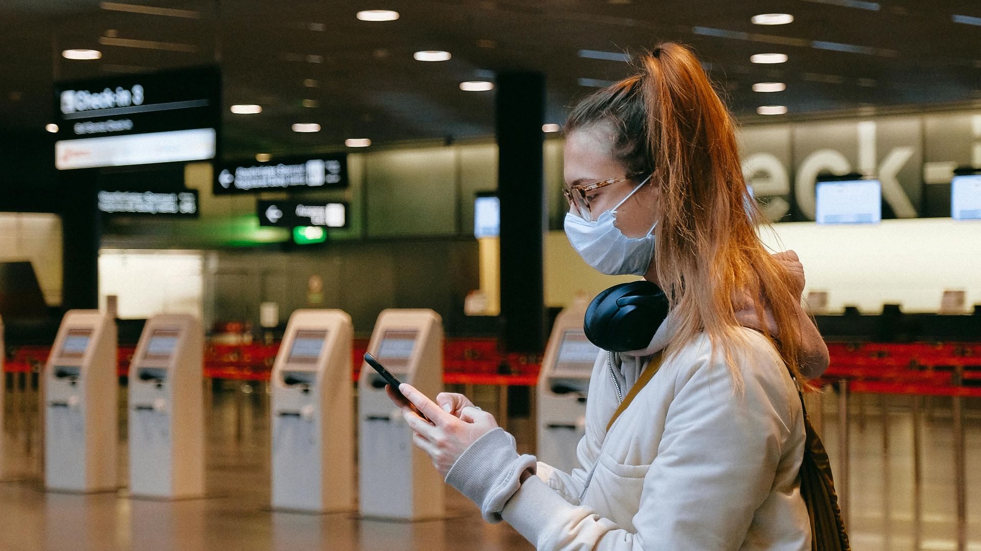 Woman wearing a mask, texting in airport terminal with signage in background.