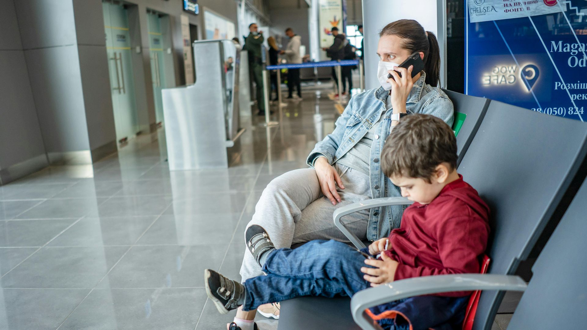 A mother and child sitting at an airport terminal. The mother is on the phone while the child is using a device.