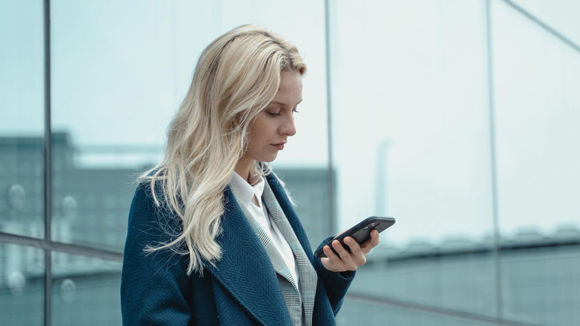 Professional woman in blue coat using smartphone outside office building.