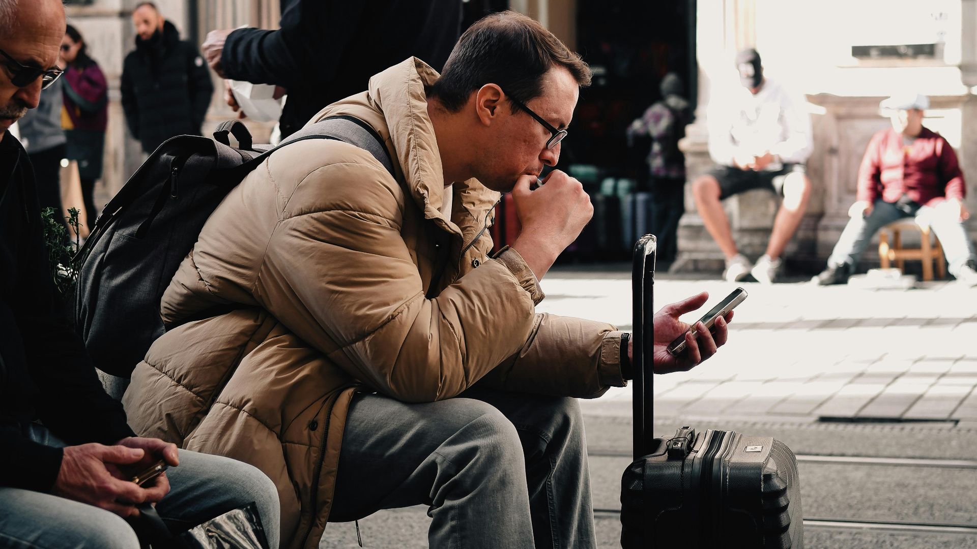 A man sitting with luggage, checking his phone in a bustling urban street scene, surrounded by people.