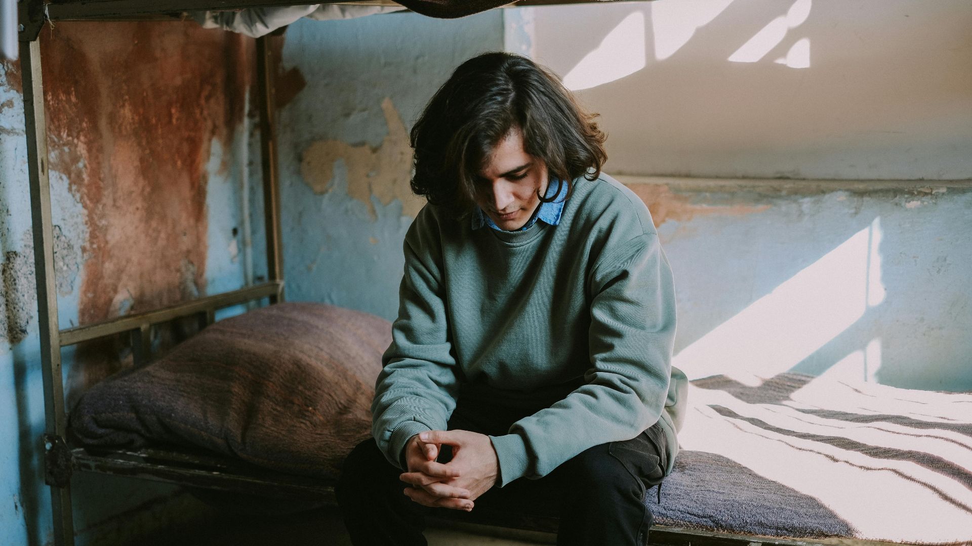 A man in a sweatshirt sits thoughtfully on a bunk bed in a rustic room with textured walls.