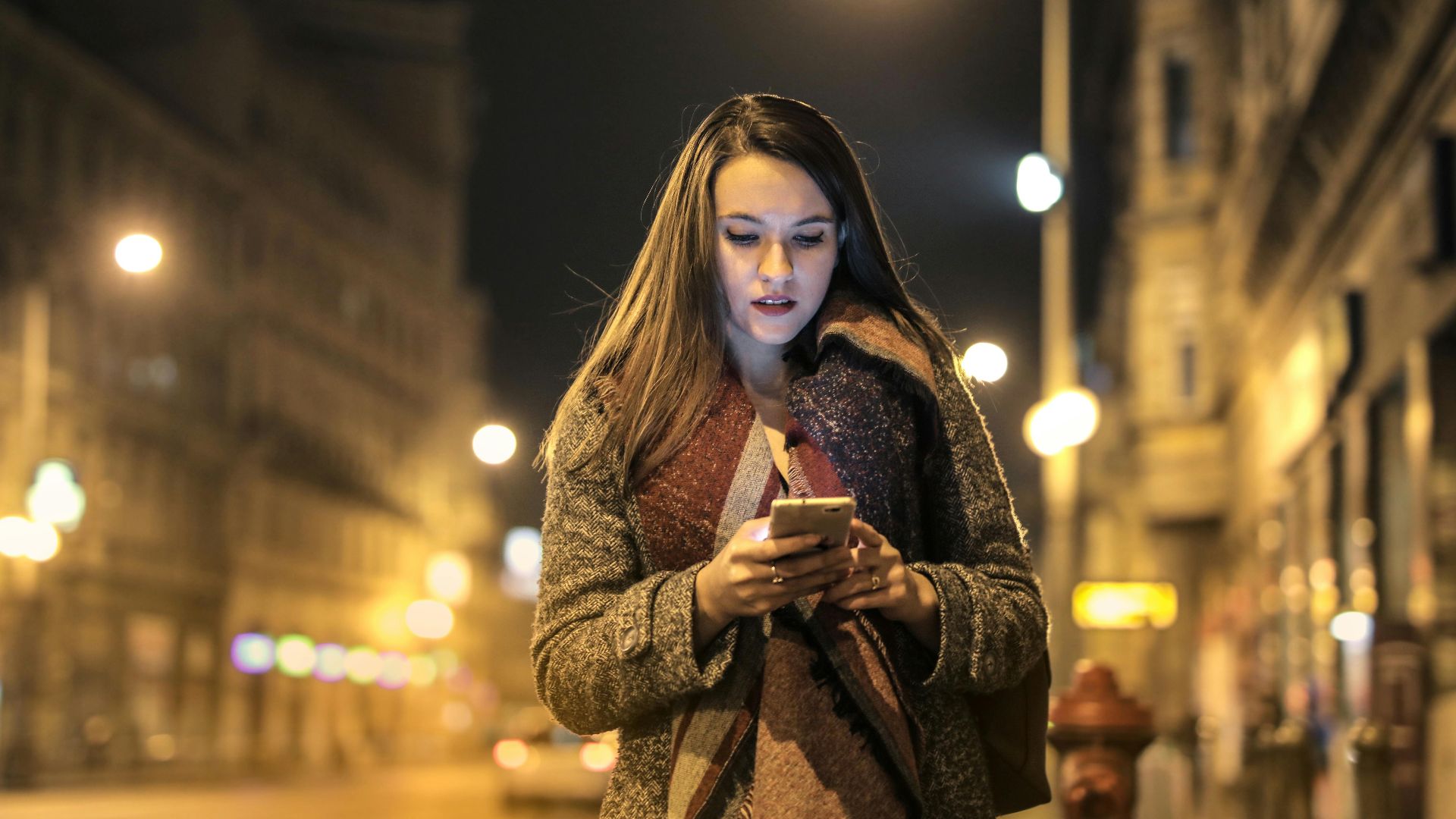 Elegant woman walks through urban street at night, engaged with her smartphone, surrounded by city lights.