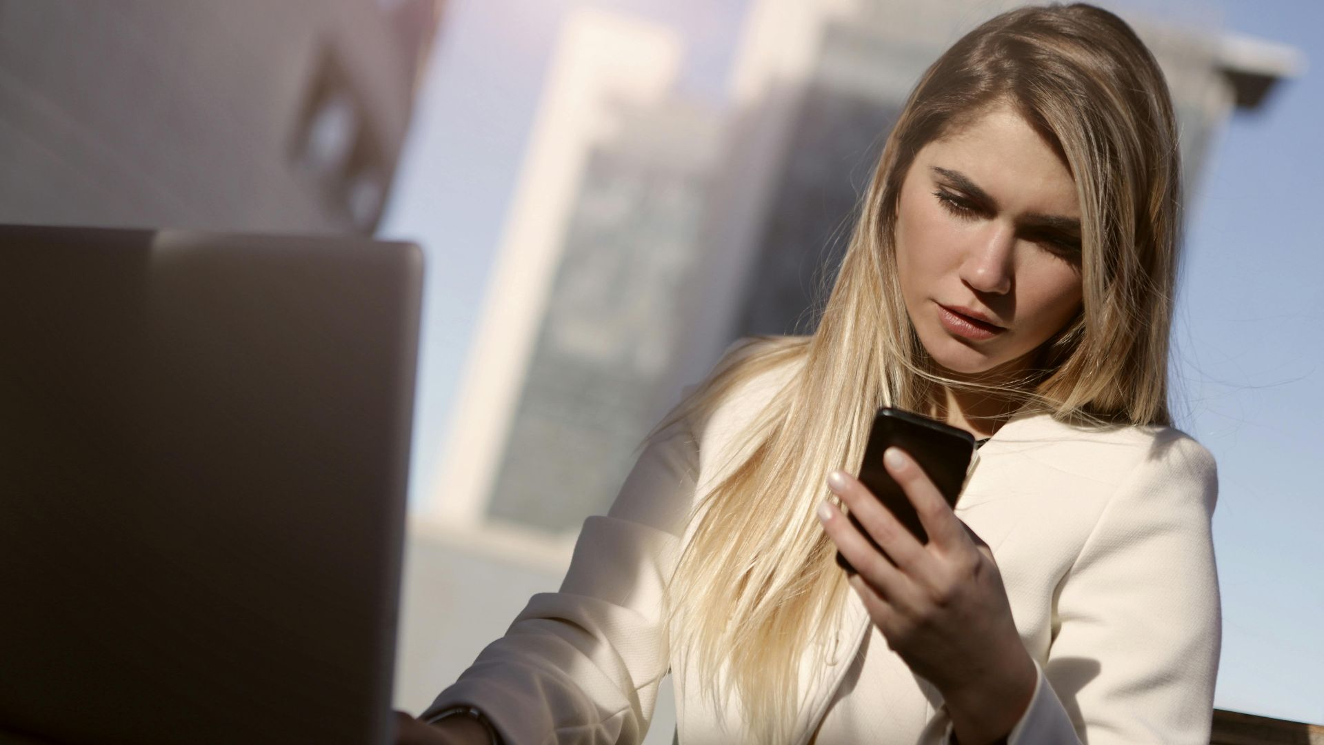 Focused businesswoman using a smartphone outdoors with laptop, modern lifestyle.