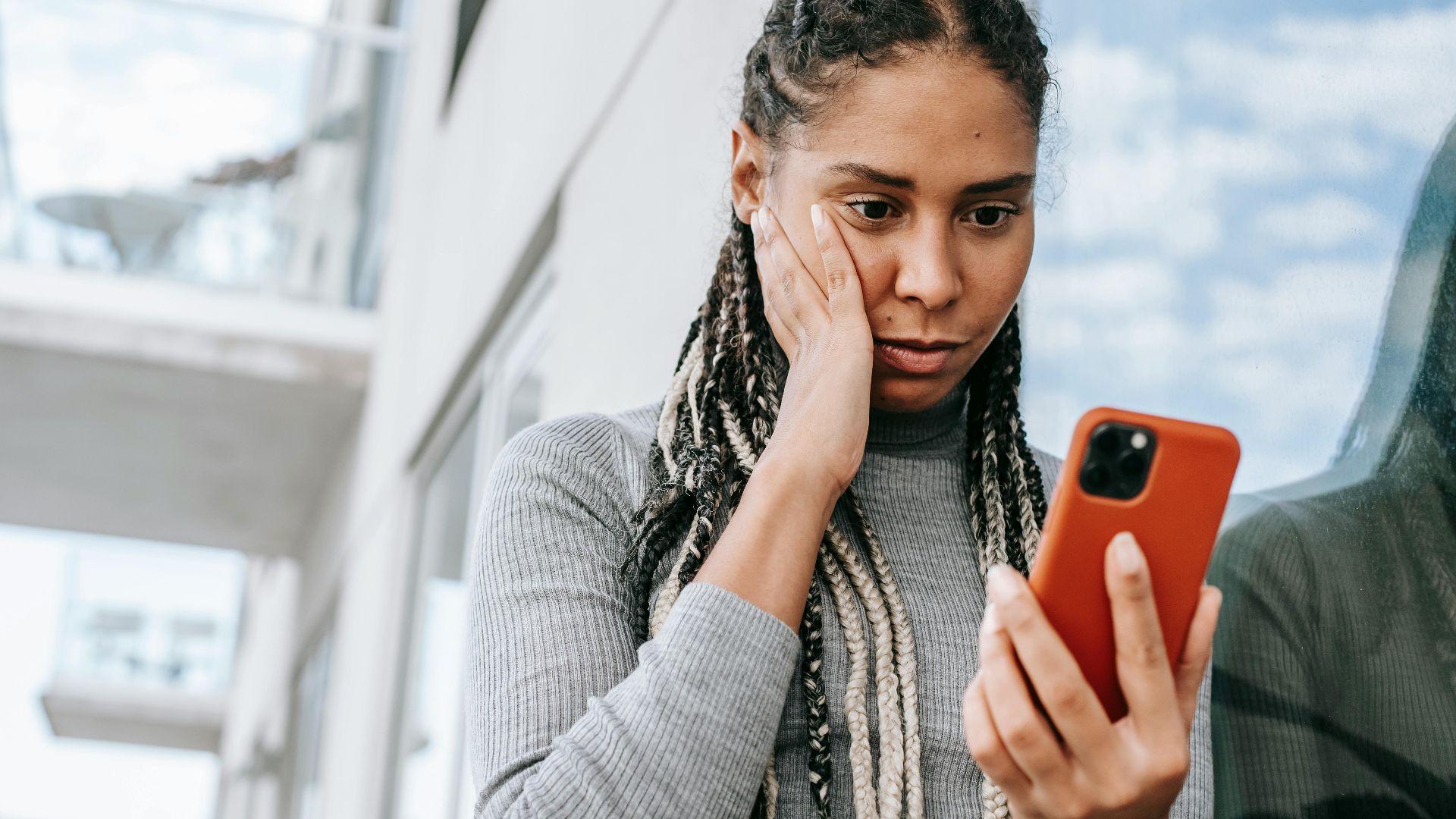 Crop serious African American female with dreadlocks touching cheek in contemplation and browsing mobile phone on street