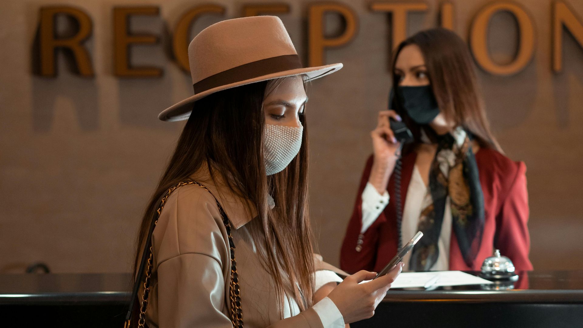 Woman in face mask at hotel reception counter checking her phone, symbolizing the new normal.