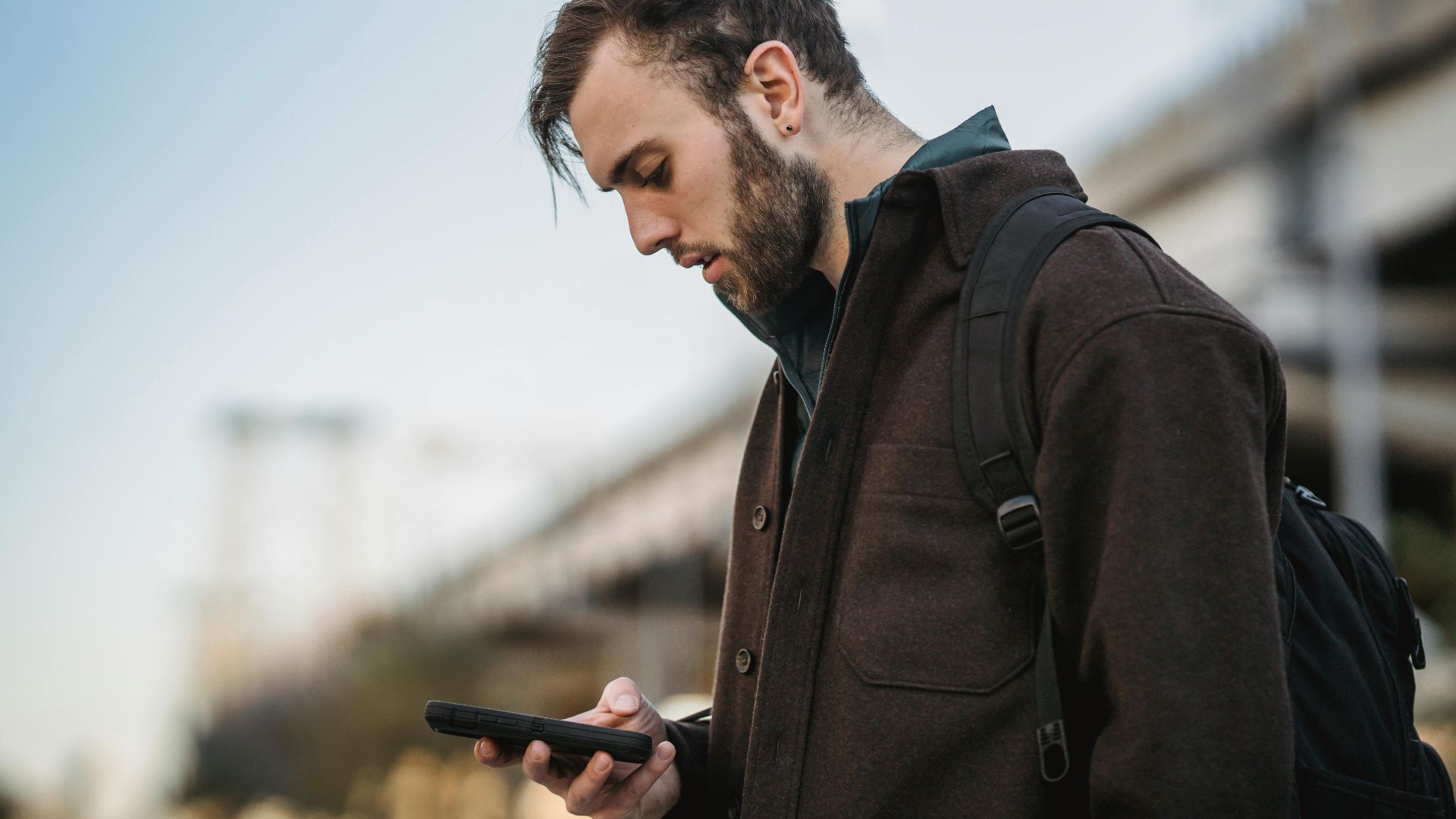 Side view of crop young unshaven male watching cellphone under light sky in town