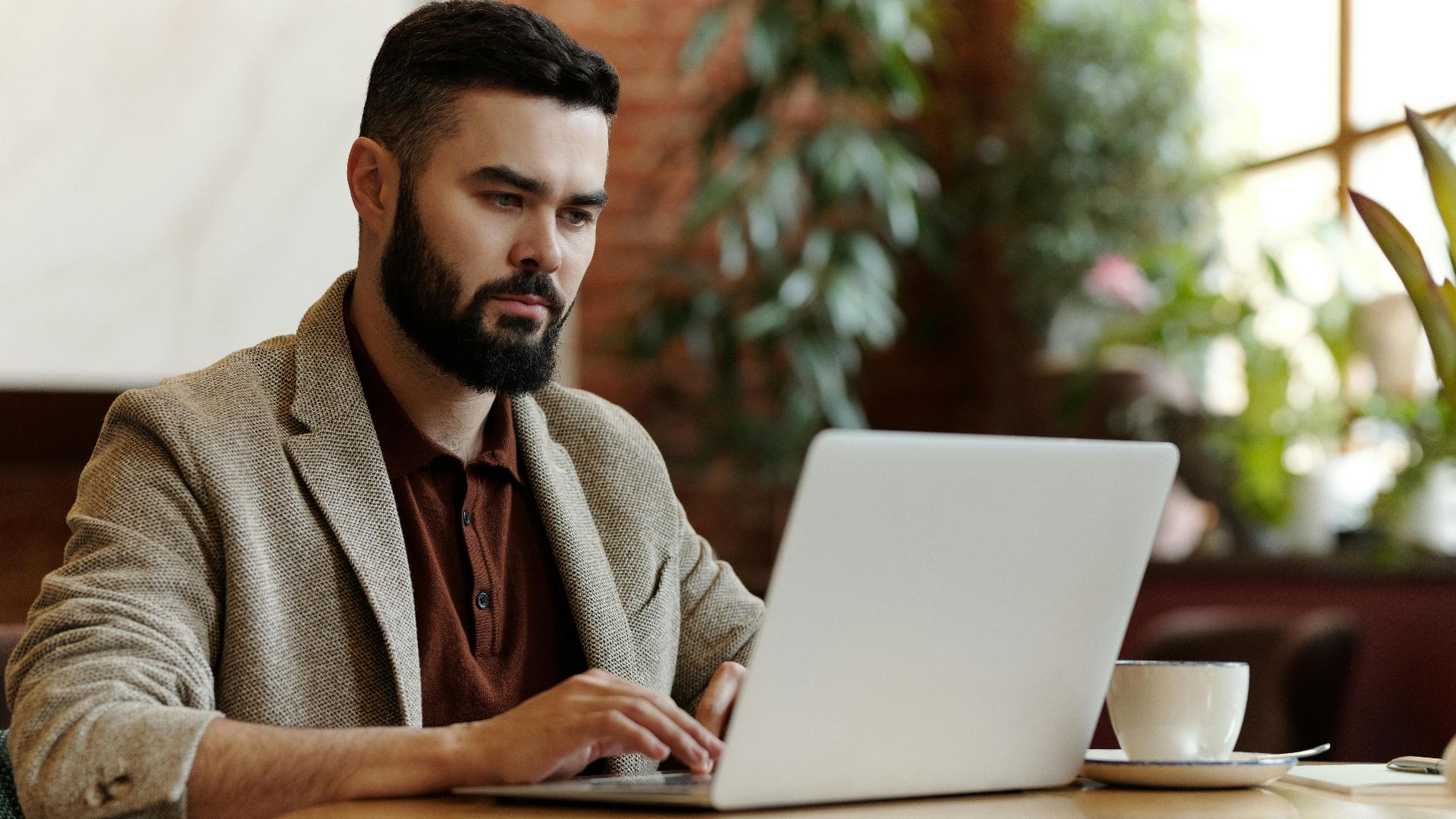 Businessman working on laptop in a cafe setting, surrounded by plants, sipping coffee.
