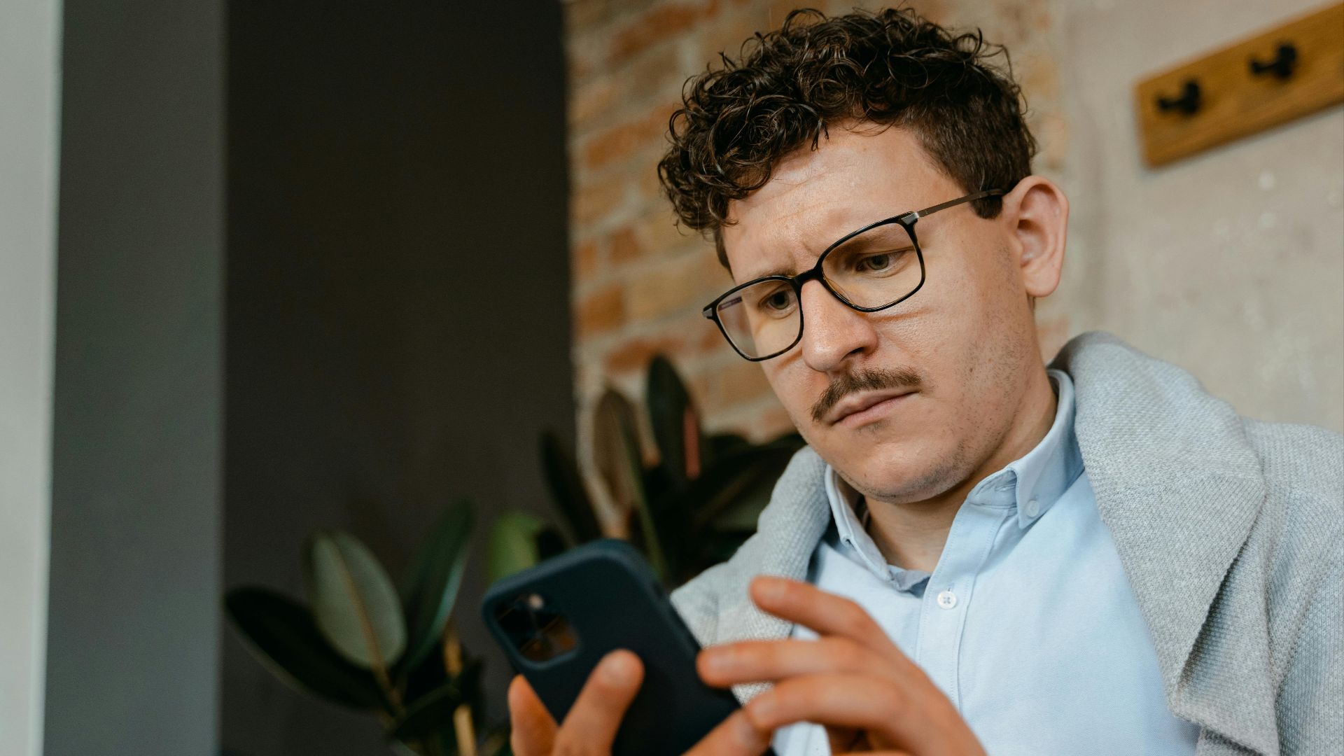 A focused man with glasses using a smartphone indoors next to a brick wall.