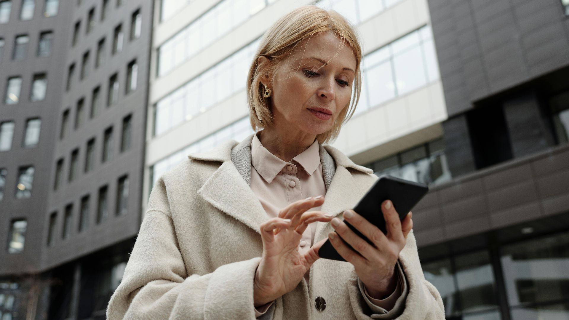 Woman in coat using smartphone in front of modern building. Professional and focused expression.