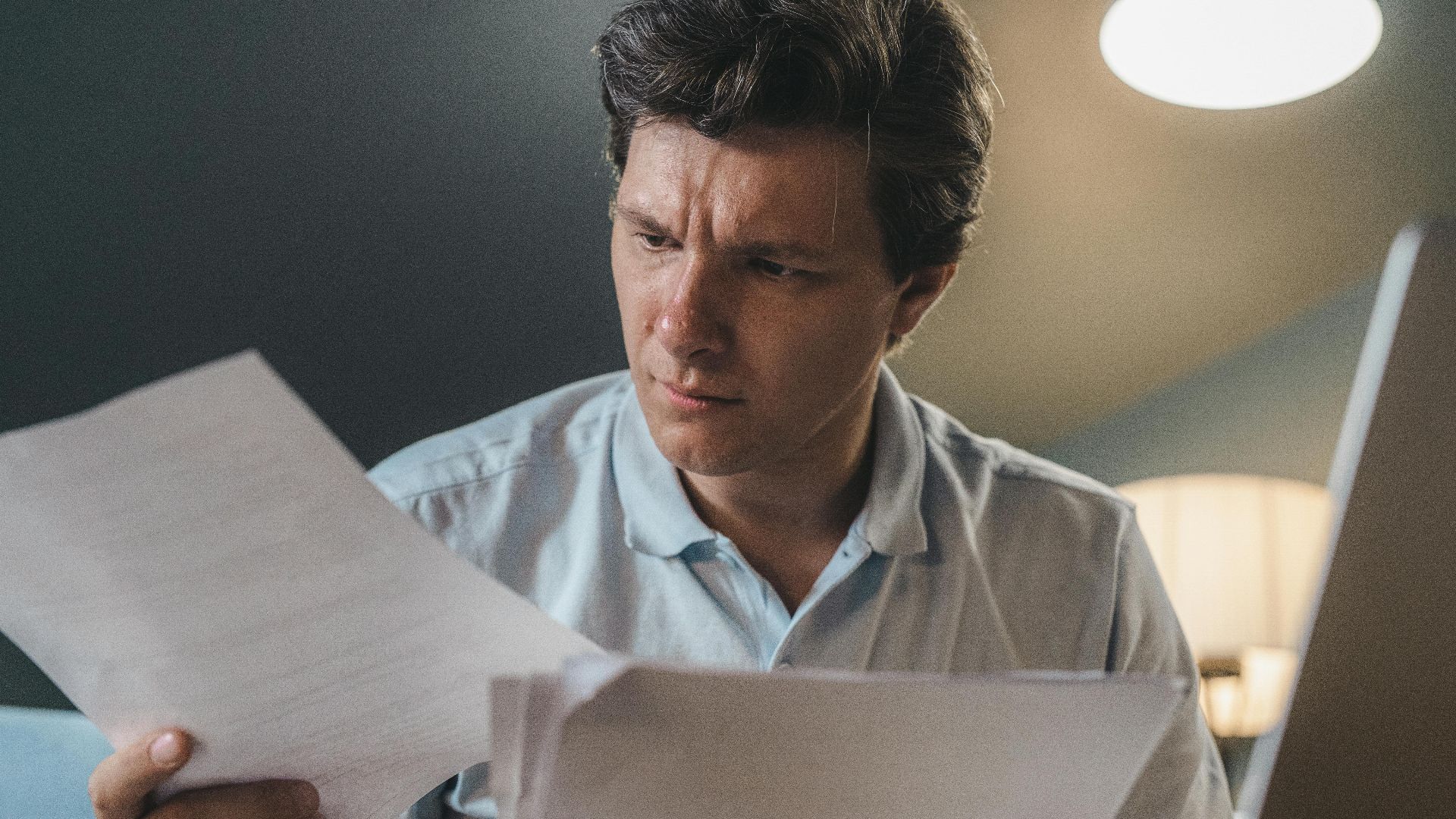 A man intently examines papers, seated indoors under warm lighting, focusing on his work.