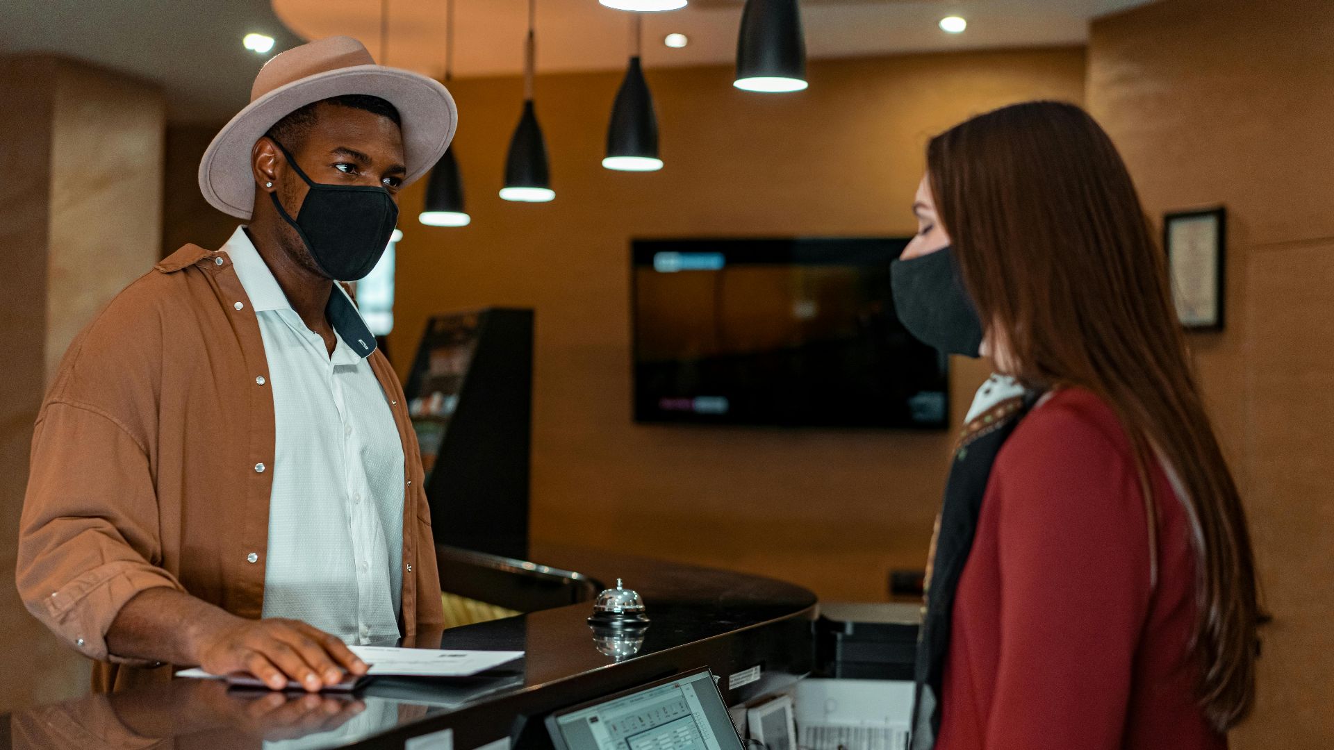 A man and woman in face masks checking in at a hotel reception, emphasizing safety protocols.