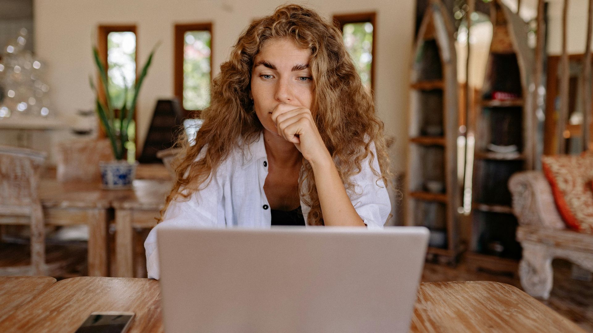 A thoughtful woman working remotely on a laptop in a cozy indoor setting.