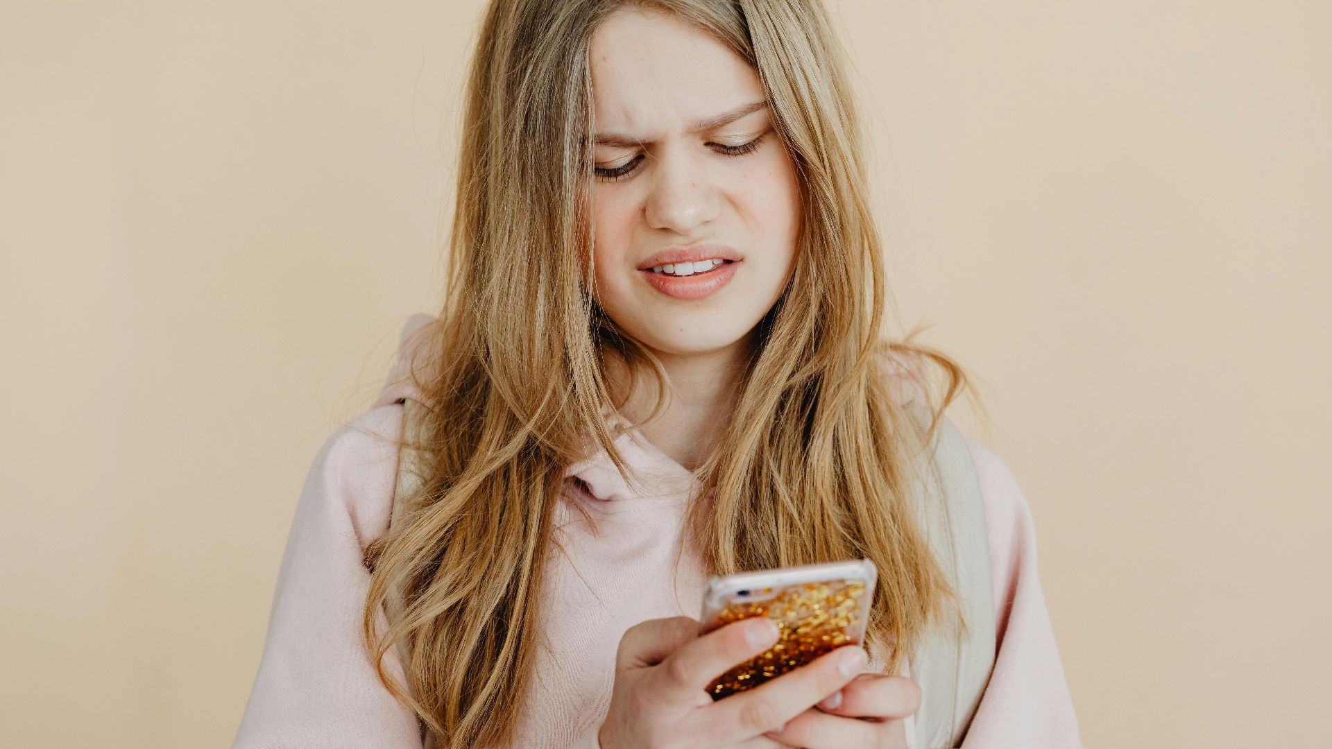 Teen girl with blonde hair in pink hoodie using smartphone, expressing curiosity indoors.
